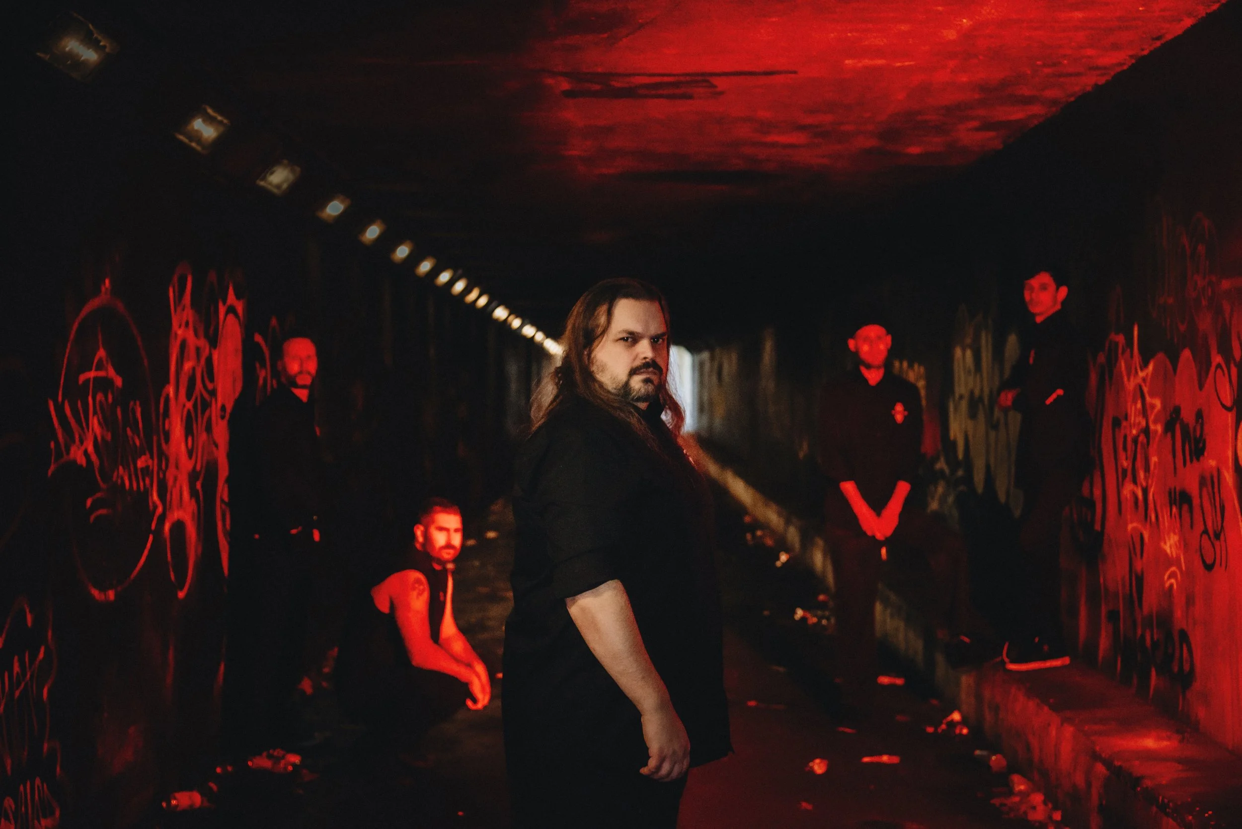 A group of five men in a dimly lit tunnel with graffiti-covered walls, illuminated by red lighting. One man with long hair and a beard stands in the foreground, looking at the camera with a serious expression.
