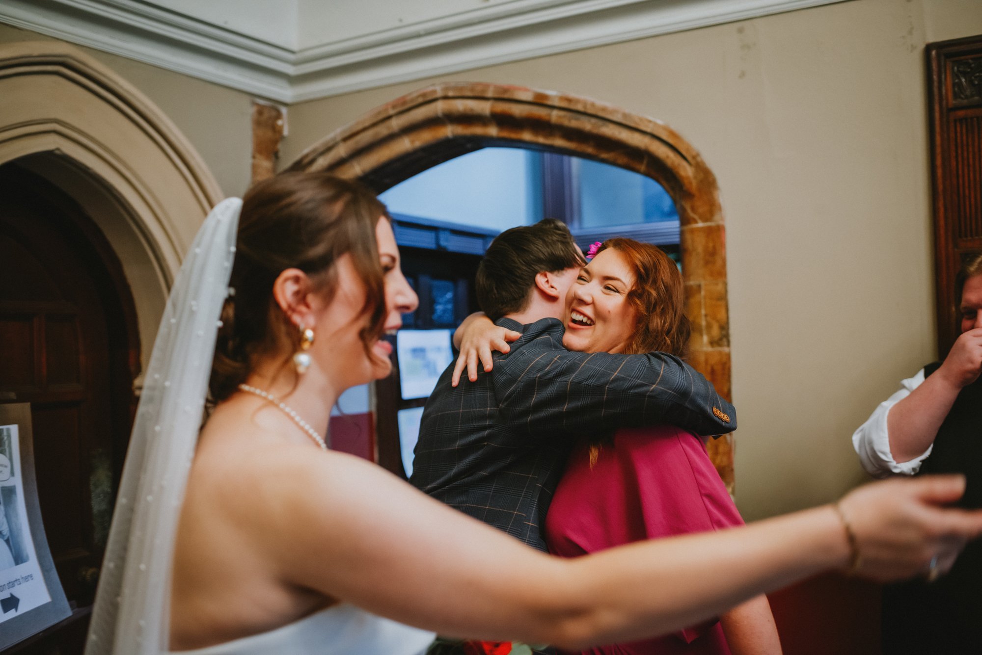 A bride in a white dress with a veil taking a selfie with a smiling woman in a pink dress and a man hugging her, celebrating at a wedding reception.