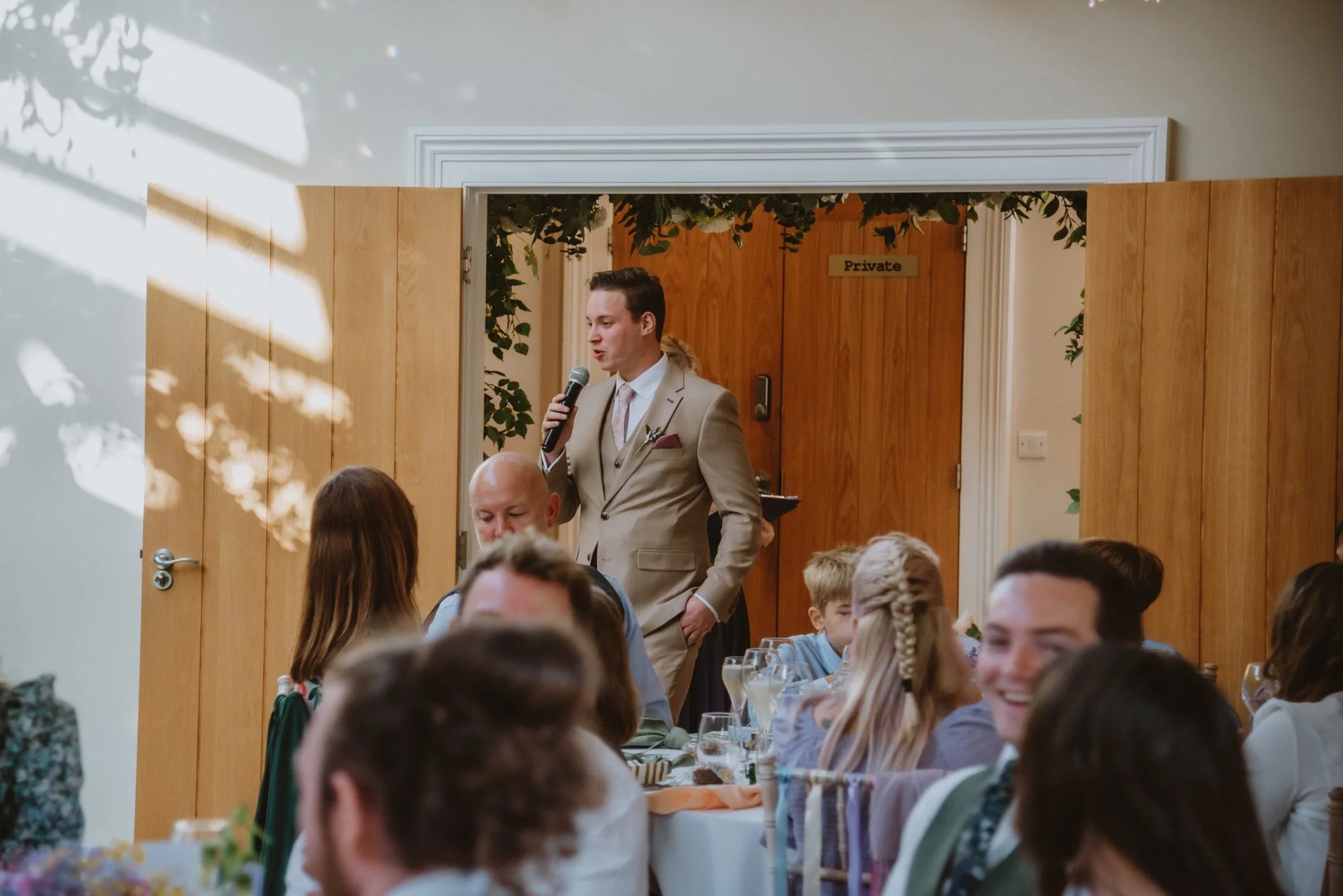 A man in a beige suit speaking into a microphone at a wedding reception, with guests seated around tables in the foreground.