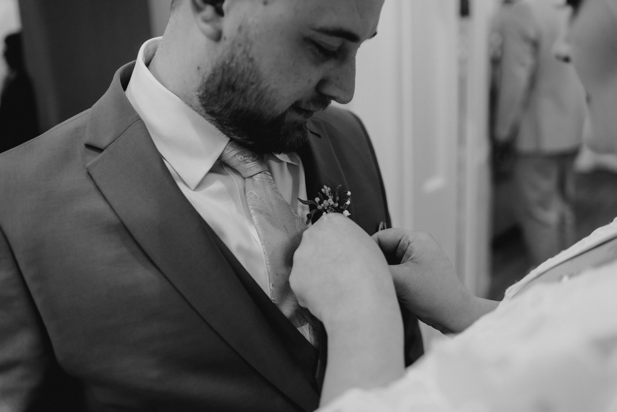 A man in a suit is having a boutonniere pinned onto his lapel by a woman at a wedding or formal event.