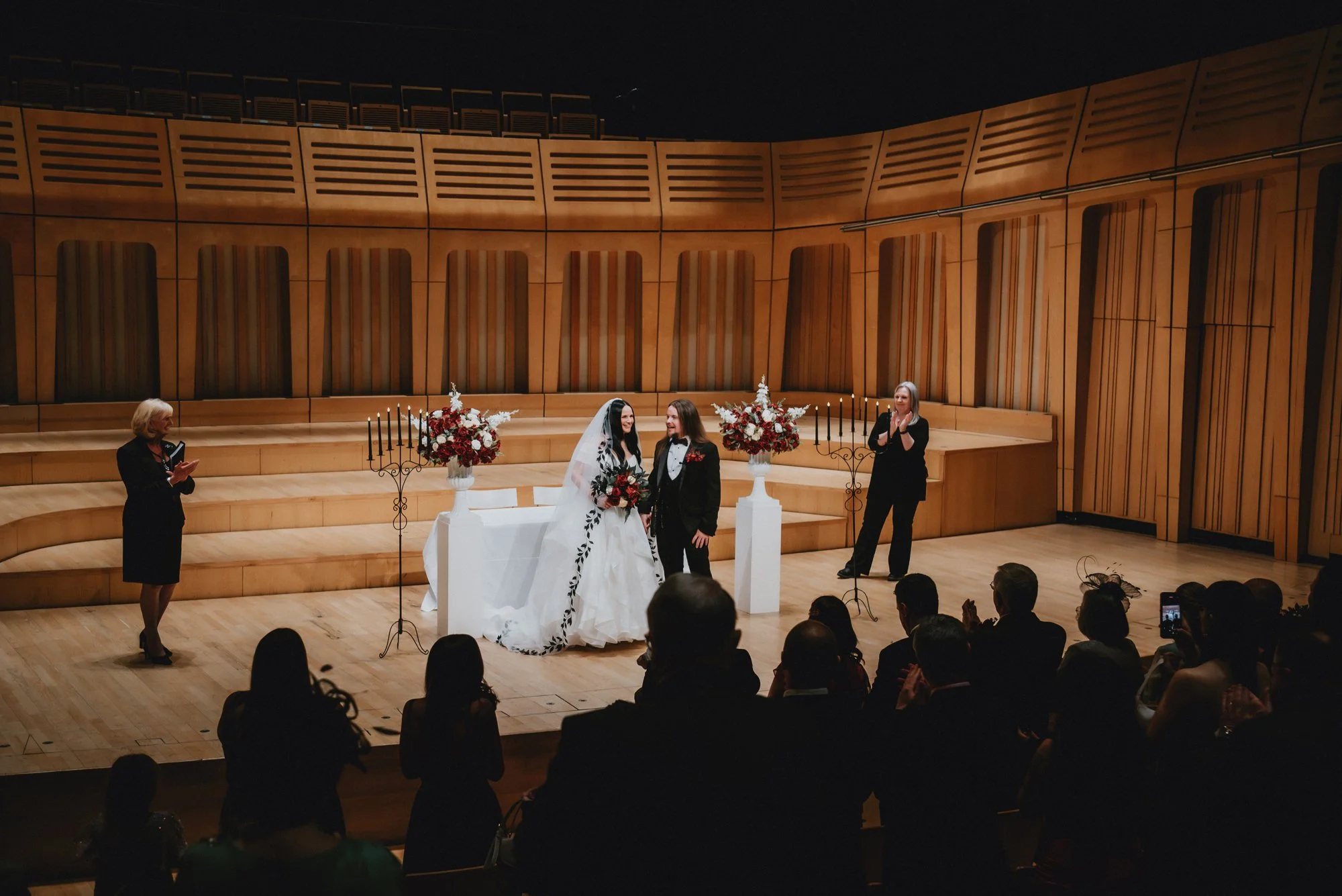 A wedding ceremony with a bride and groom on stage, surrounded by officiants and guests in an auditorium.