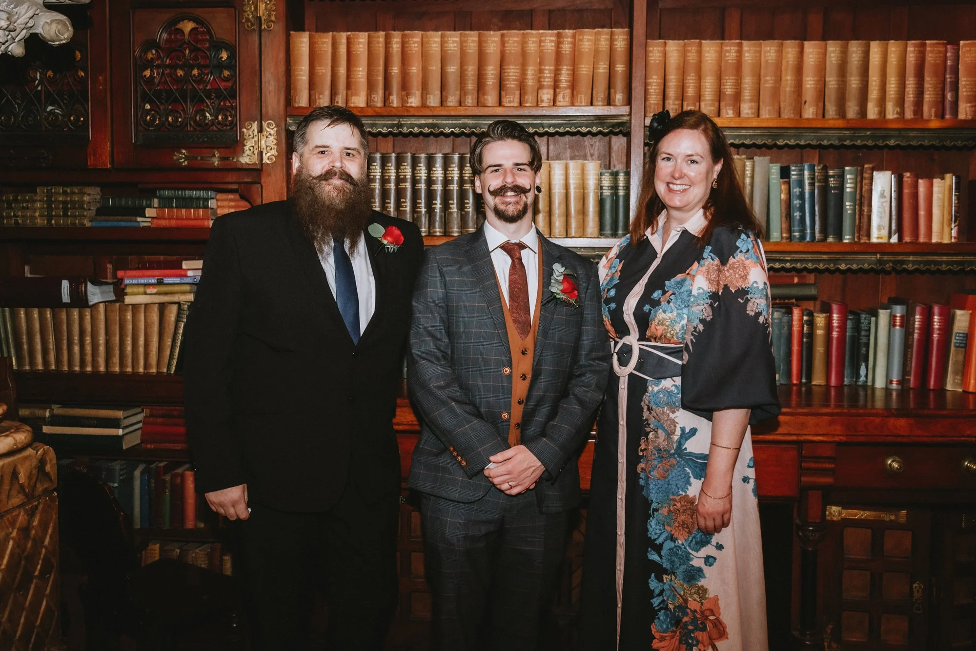 Three people standing together in front of a wooden bookshelf filled with books. The man on the left has a beard and mustache, is wearing a black suit with a blue tie and a red flower pin. The man in the middle has short hair and a beard, is dressed 