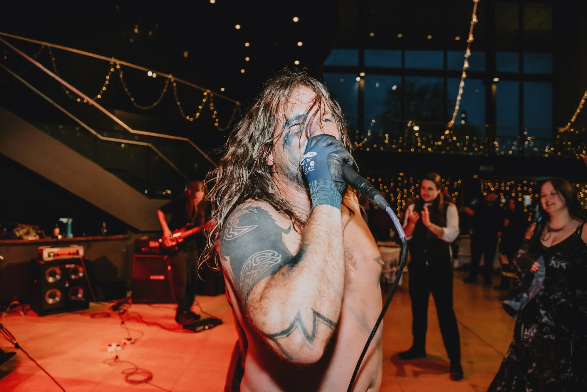 A shirtless male singer with long, wavy hair and tattoos on his arm, holding a microphone and singing at a party or concert. Several people are dancing and enjoying the music in the background, with string lights and a balcony visible.