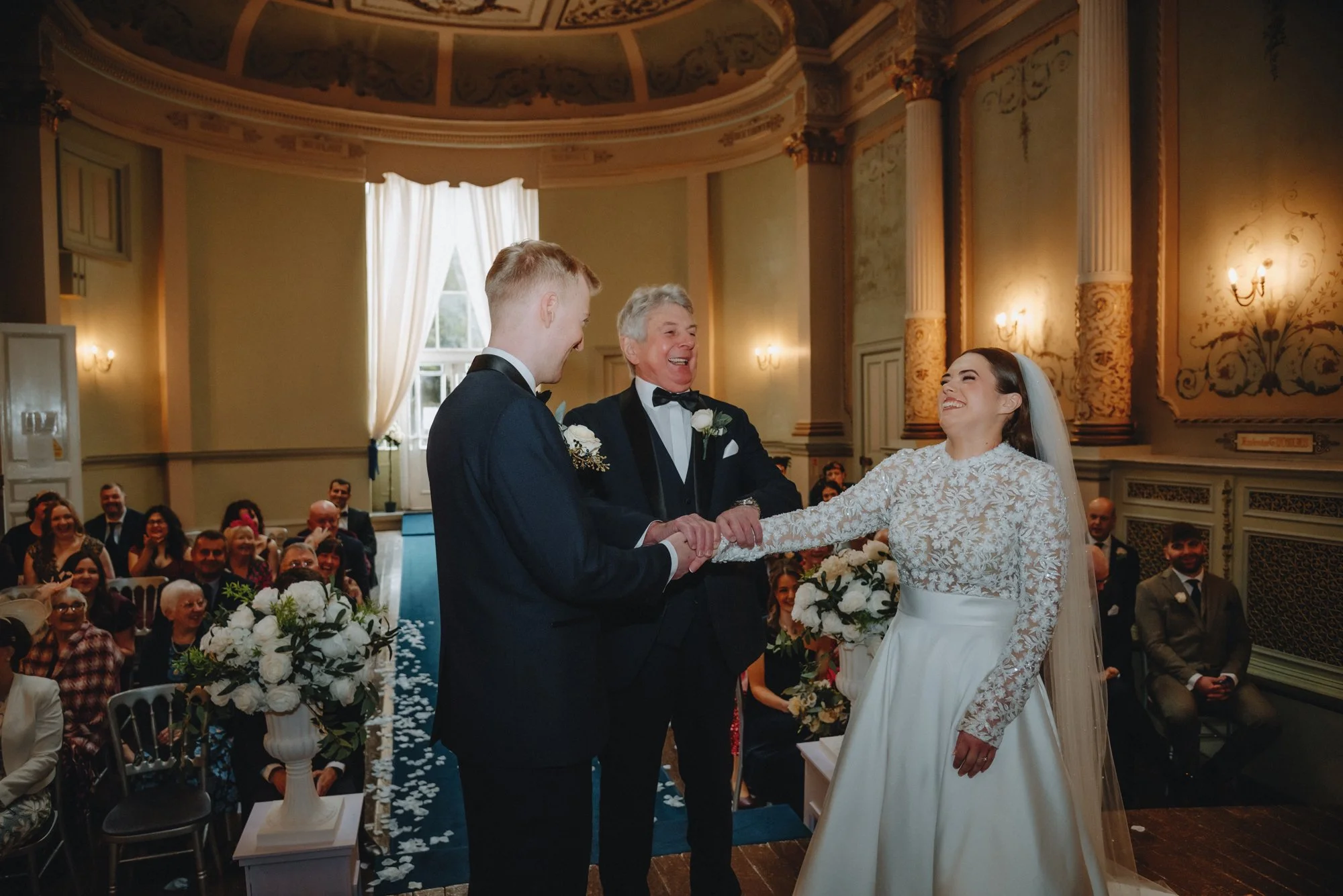 A bride and groom holding hands and smiling during their wedding ceremony, standing between an officiant and a group of guests seated in an elegant hall with historic decor and floral arrangements.