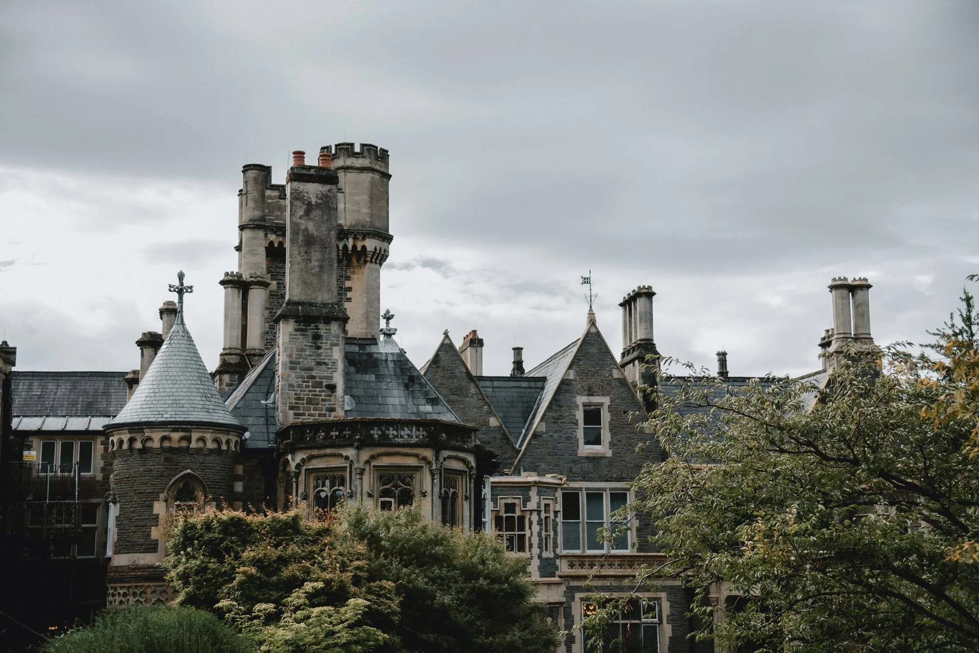 Historic stone mansion with multiple turrets, chimneys, and ornate windows, partially obscured by trees, under a cloudy sky.
