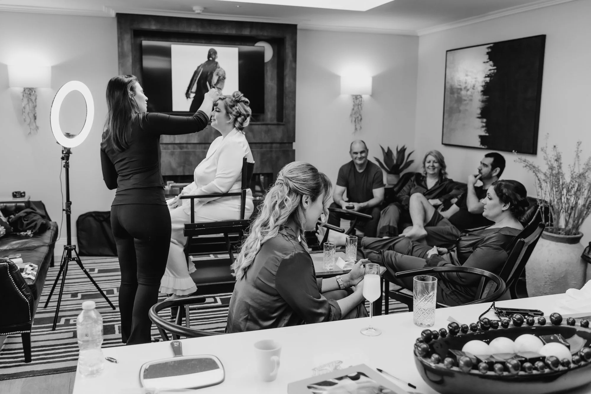 People in a living room, with one woman sitting on a chair having her makeup done by another woman, while others sit on the couch and chairs watching. The scene includes a ring light, framed artwork, and various drinks on a table.