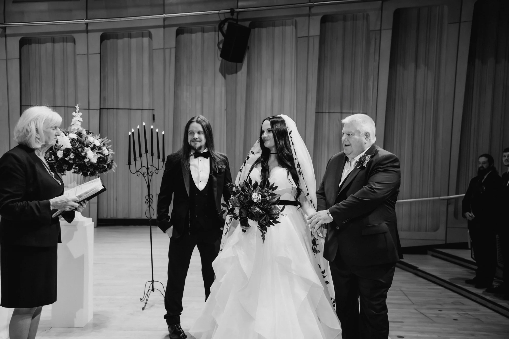 A wedding ceremony with a bride holding a bouquet, a groom, and two other individuals standing with them, inside a decorated hall.