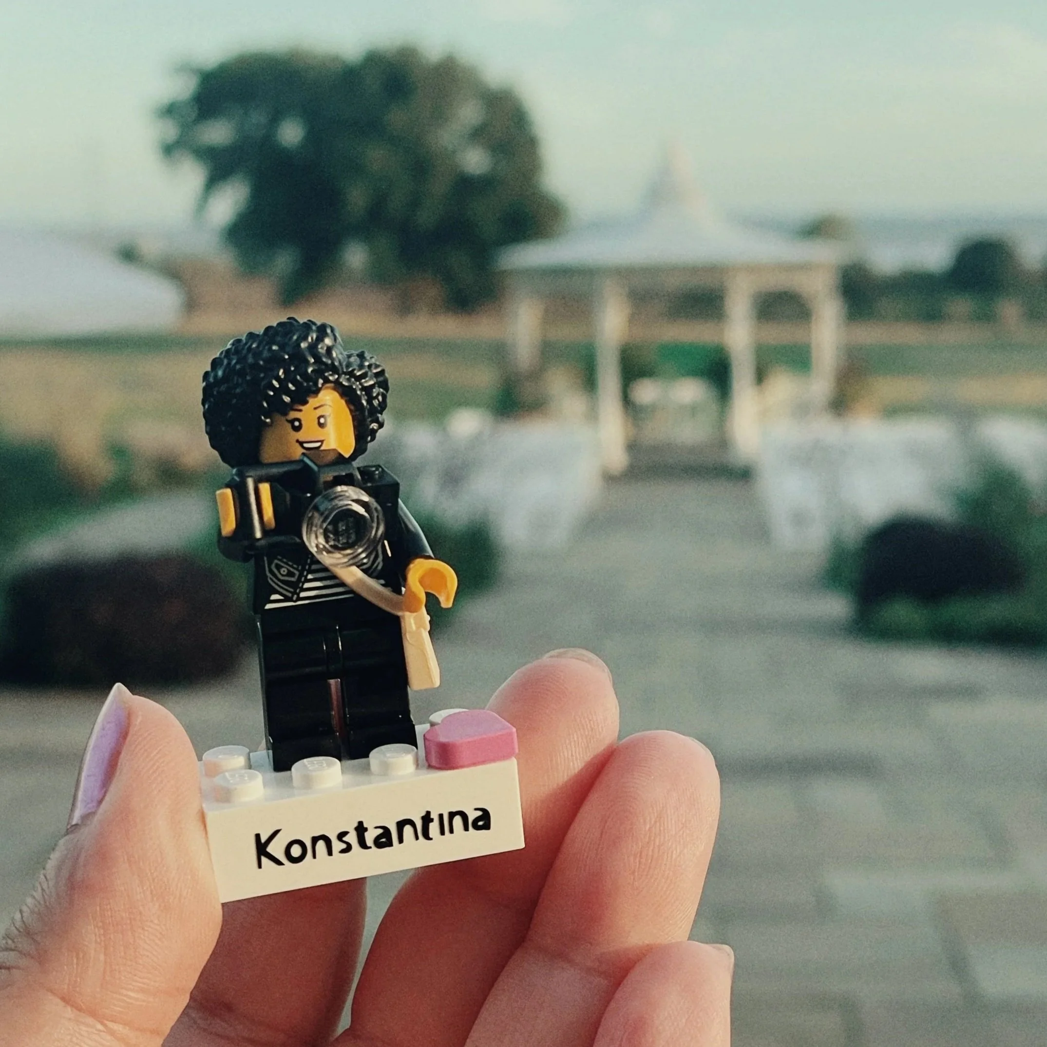 Lego figure of a woman with curly hair, holding a camera, standing on a white brick with the name 'KONSTANTINA' written on it, with a park or garden with a gazebo and trees in the background.
