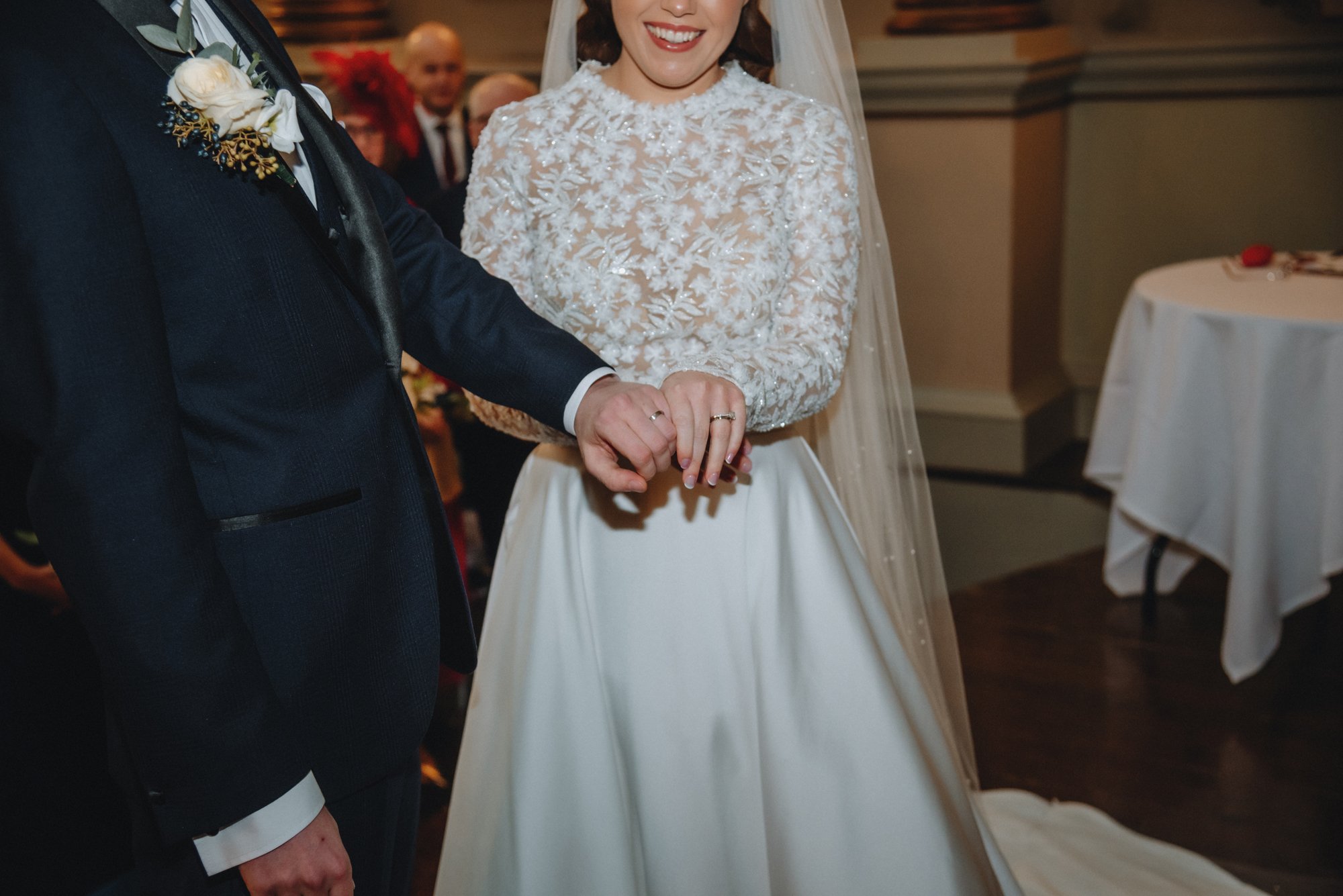 Bride and groom holding hands during wedding ceremony, with the bride in a white gown with lace detail and the groom in a dark suit with a boutonniere.