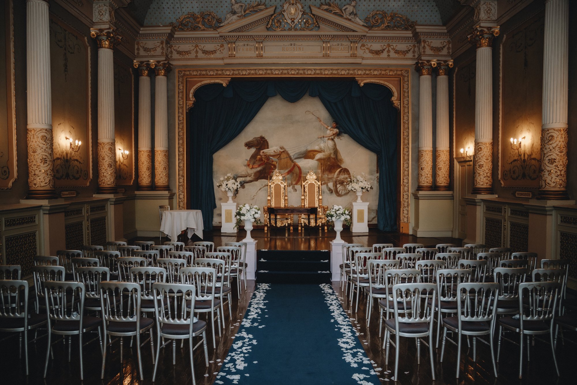 An elegant indoor wedding venue with rows of chairs facing a decorated stage, a blue carpet aisle with white petal confetti, a large art mural of a chariot pulled by horses, floral arrangements, and ornate gold and white decor.