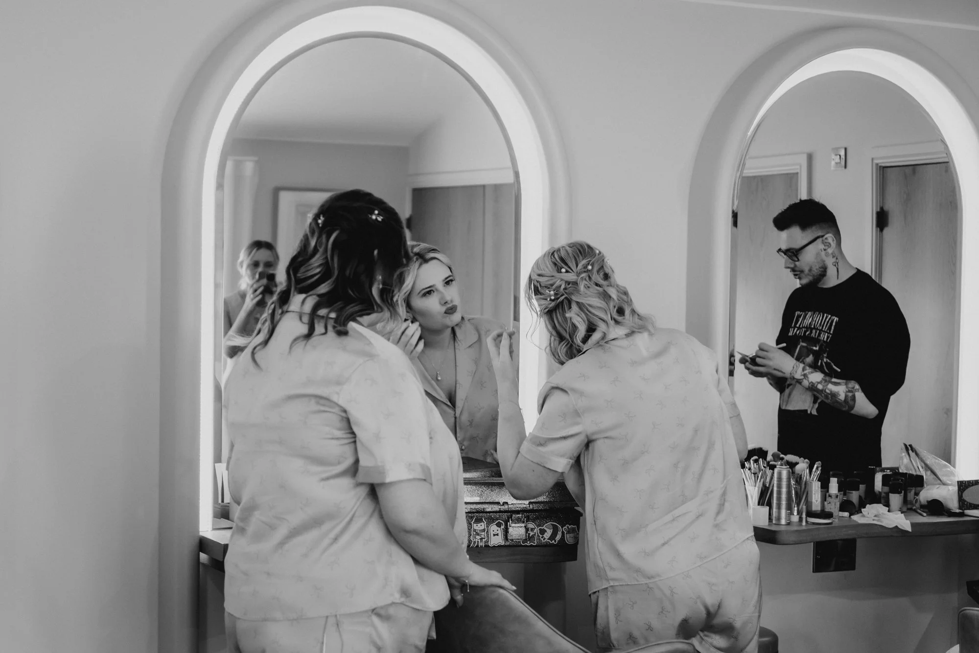 A woman applying makeup in front of a mirror while two other women assist her. A man with tattoos is looking at his phone nearby. The scene appears to be in a dressing room or backstage area.