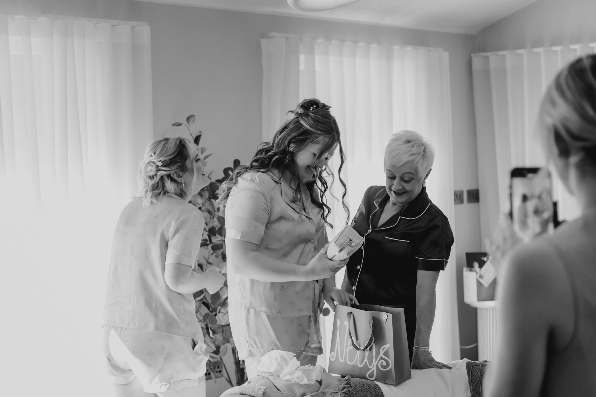 Four women in pajamas are gathered around a bed, opening gifts and smiling. One woman is holding a gift bag with "Nay's" written on it. The room has sheer curtains and potted plants in the background.