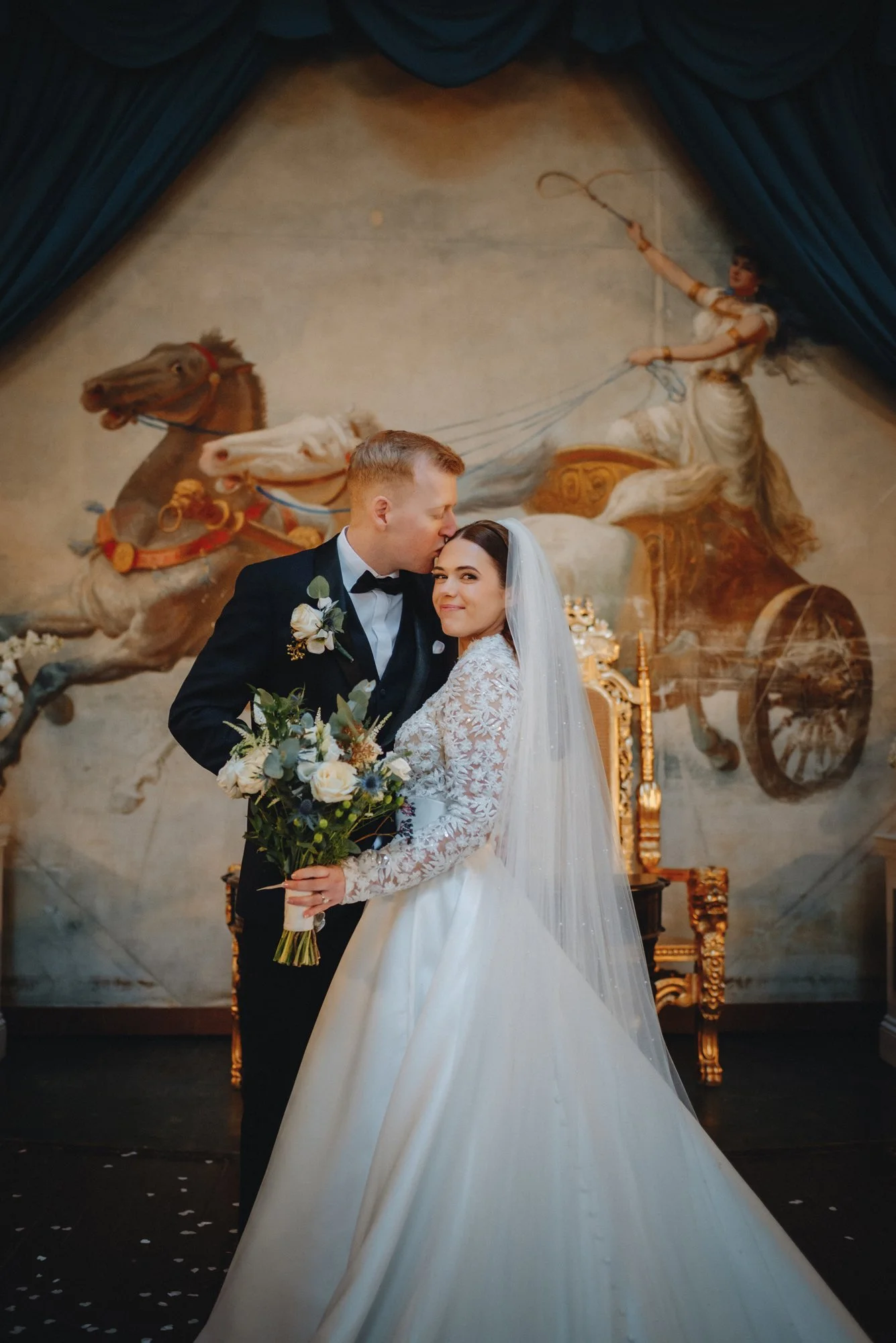 A newlywed couple, with the groom kissing the bride on the forehead, standing in front of a wall mural depicting a chariot race with a female charioteer. The bride is holding a bouquet of white and blue flowers and is smiling.