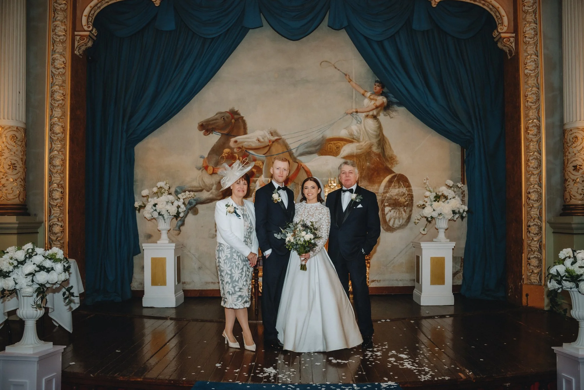 A wedding party standing on a stage with a painted backdrop of a chariot pulled by horses.