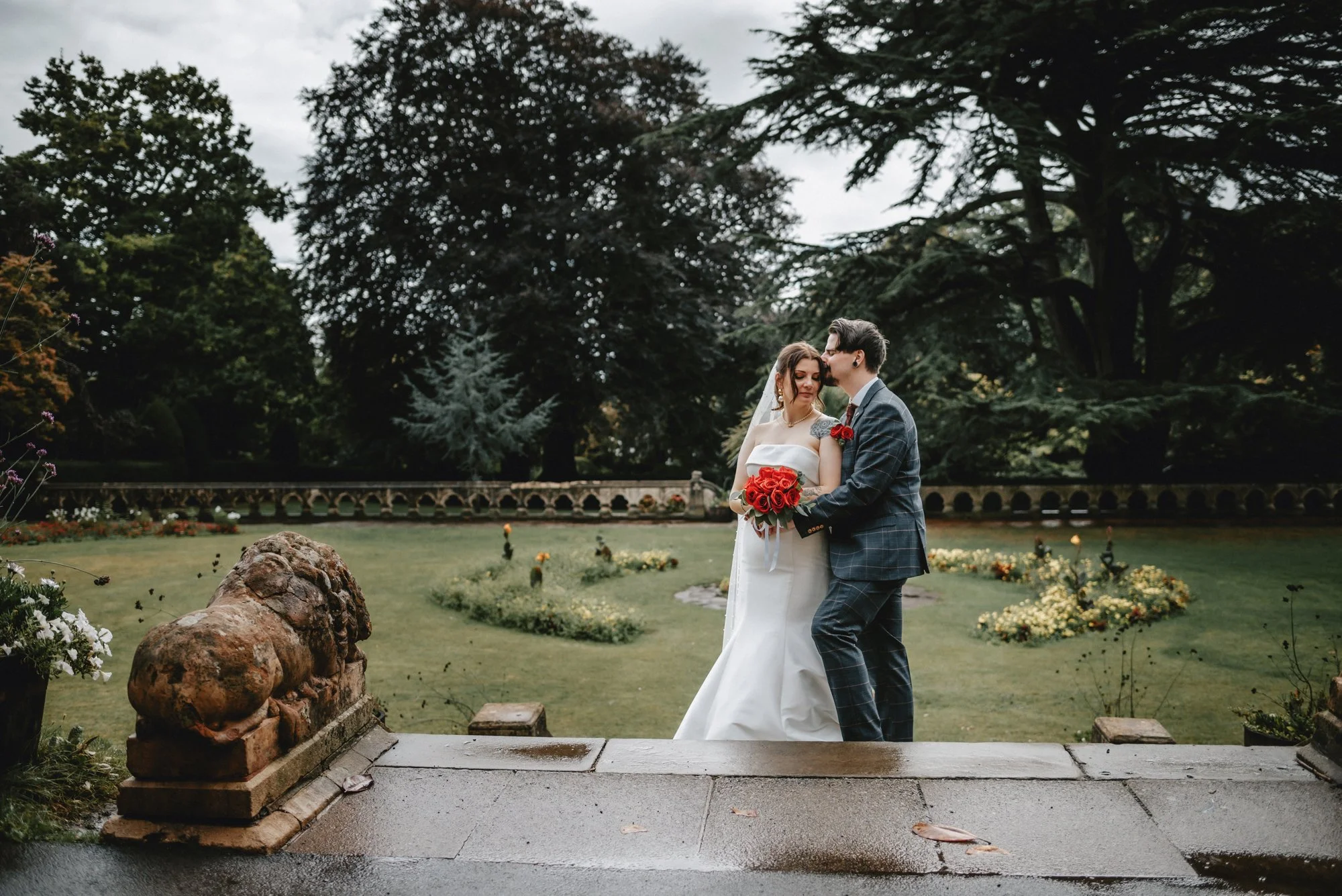 A bride and groom standing in a garden, embracing each other. The bride holds a bouquet of red roses, and the groom wears a checkered suit with a red rose boutonniere. The garden has manicured lawns, flower beds, trees, and a stone balustrade in the 