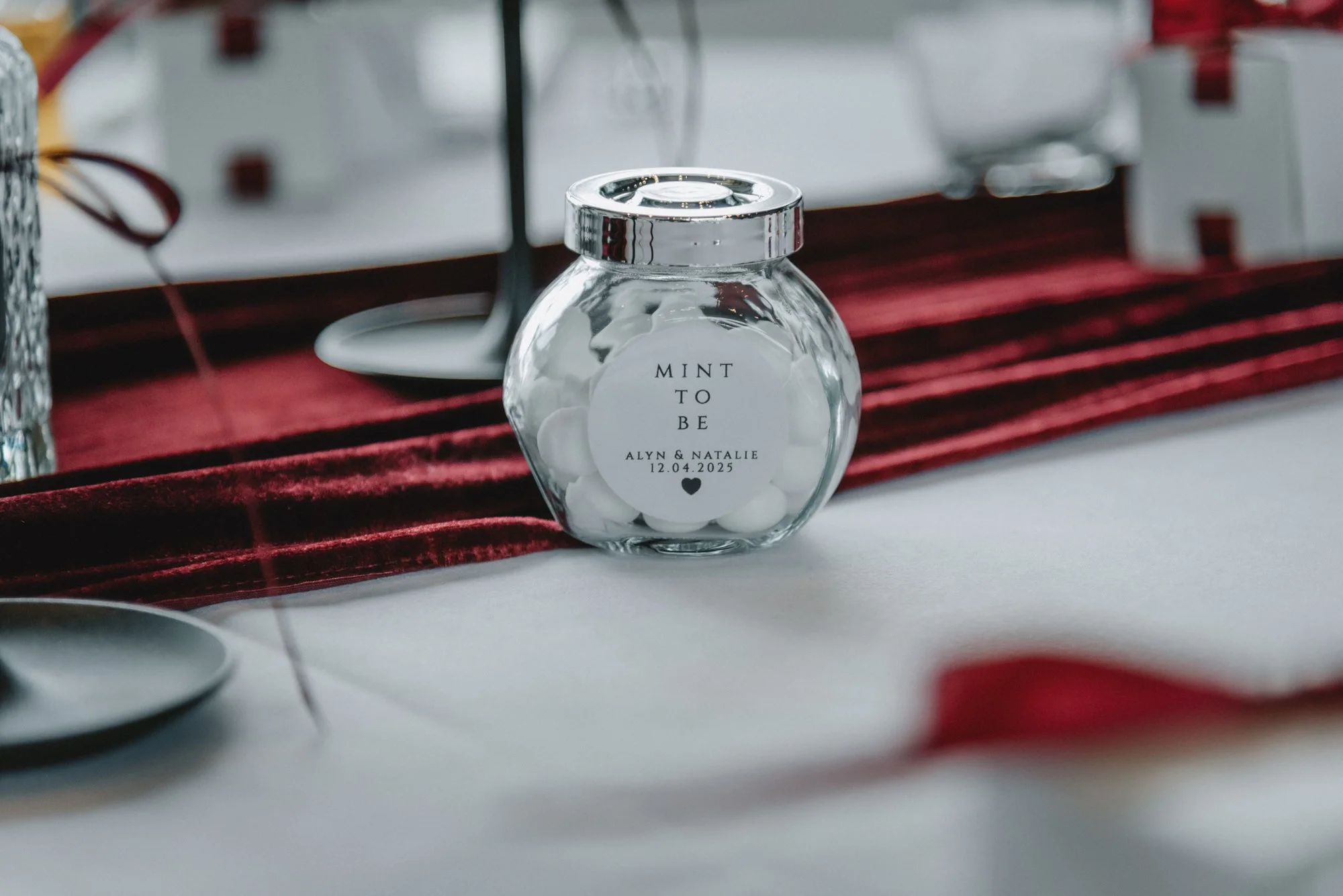 A small glass jar with a silver lid and a label that reads 'MINT TO BE ALYN & NATALIE 12.04.2025'.