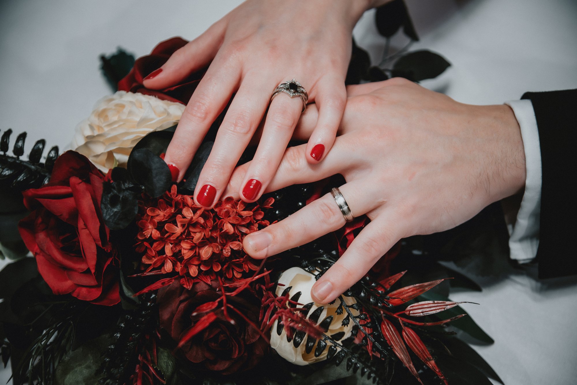 Two hands with wedding rings resting on a bouquet of red, black, and white flowers and foliage.