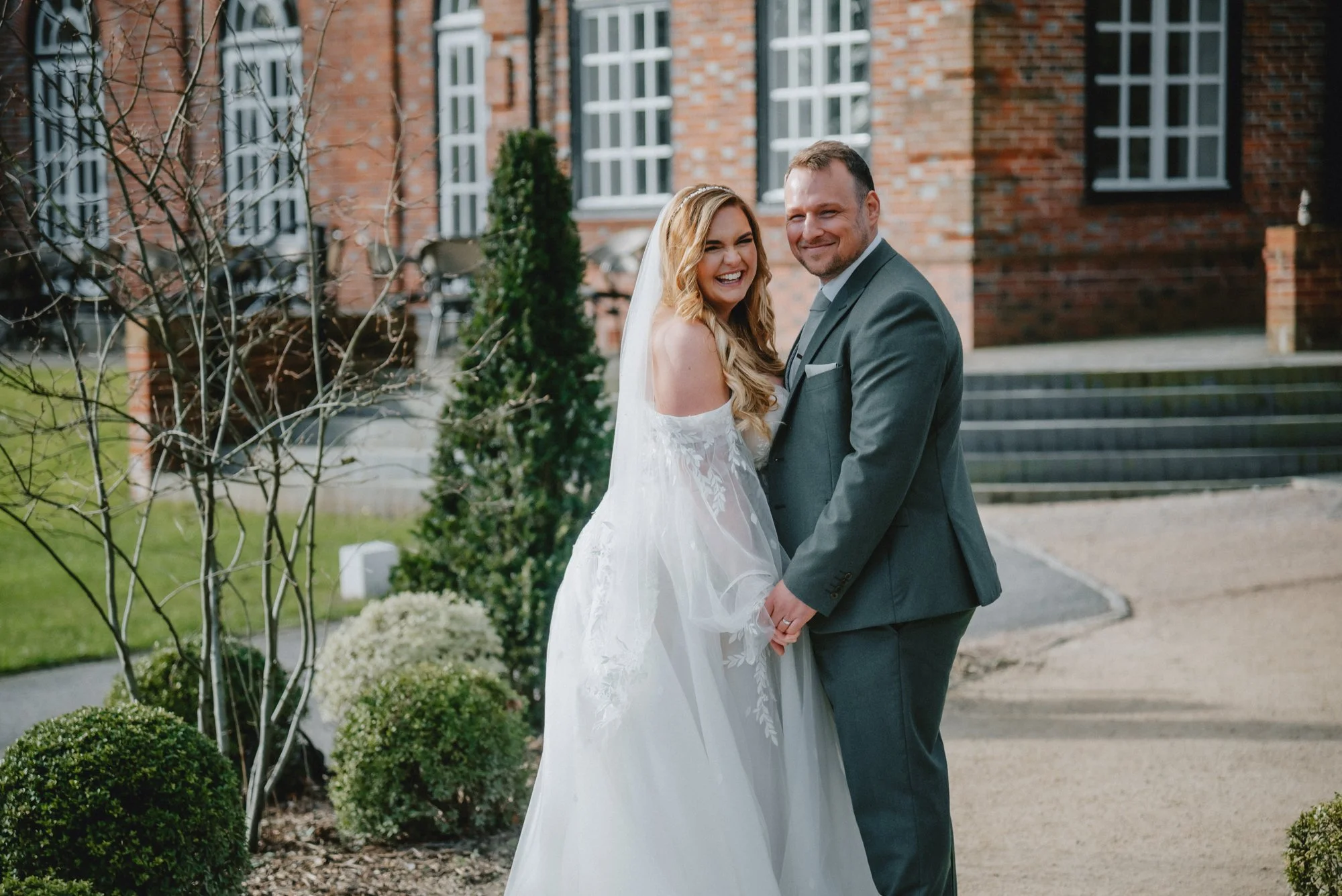 Happy bride and groom in wedding attire holding hands and smiling outdoors in front of a brick building with stairs.