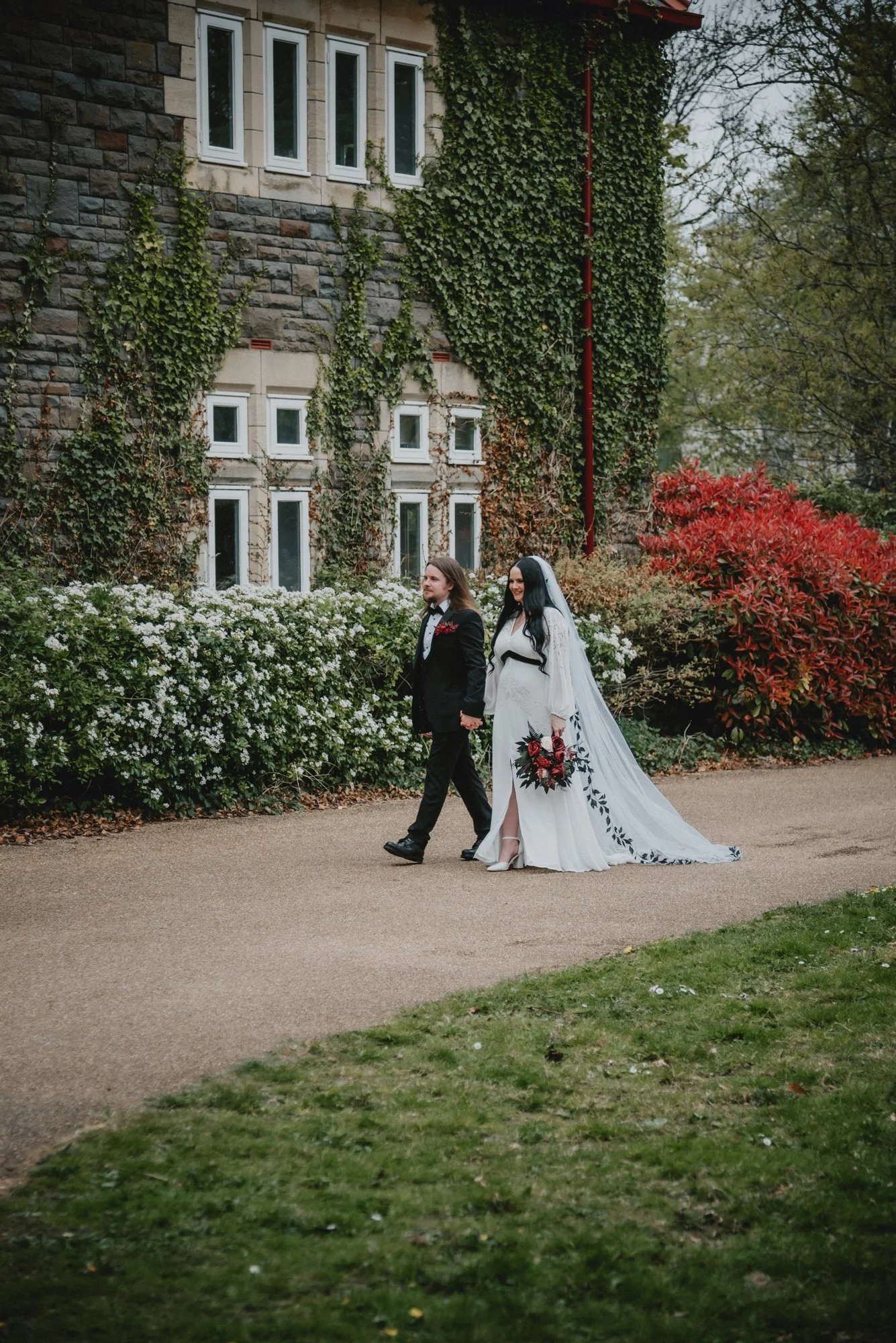 Bride and groom walking outdoors, holding hands, surrounded by lush greenery and colorful bushes, in front of a stone building with ivy-covered walls.