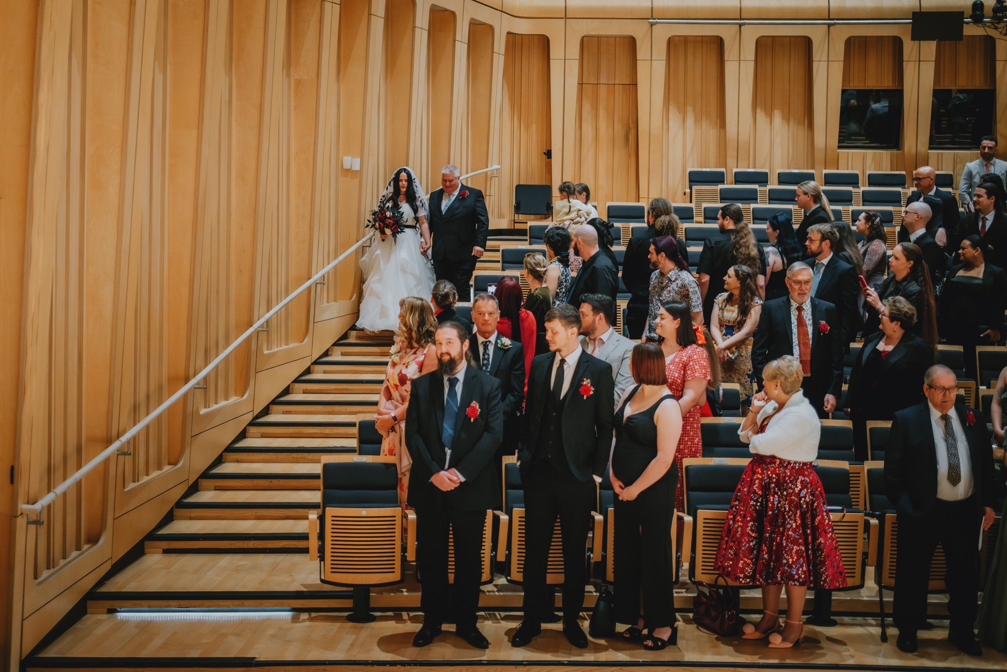 A bride walking down the stairs with her father during a wedding ceremony, with guests standing and seated in a wooden auditorium.