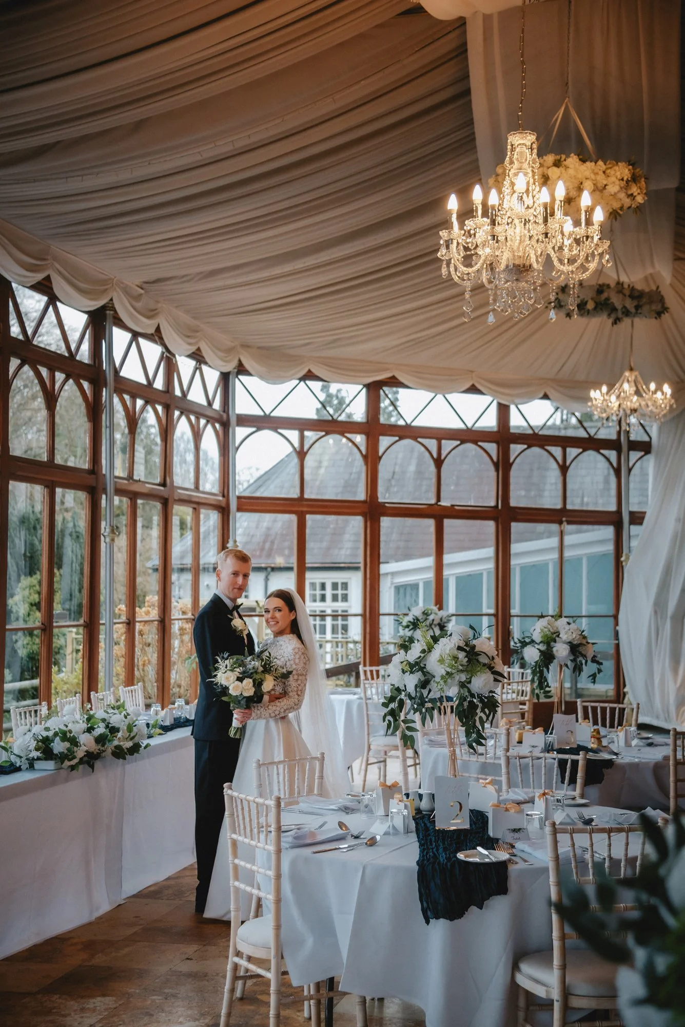 A bride and groom standing together inside a decorated wedding reception area, holding a bouquet, surrounded by round tables with white tablecloths, floral centerpieces, and table numbers.
