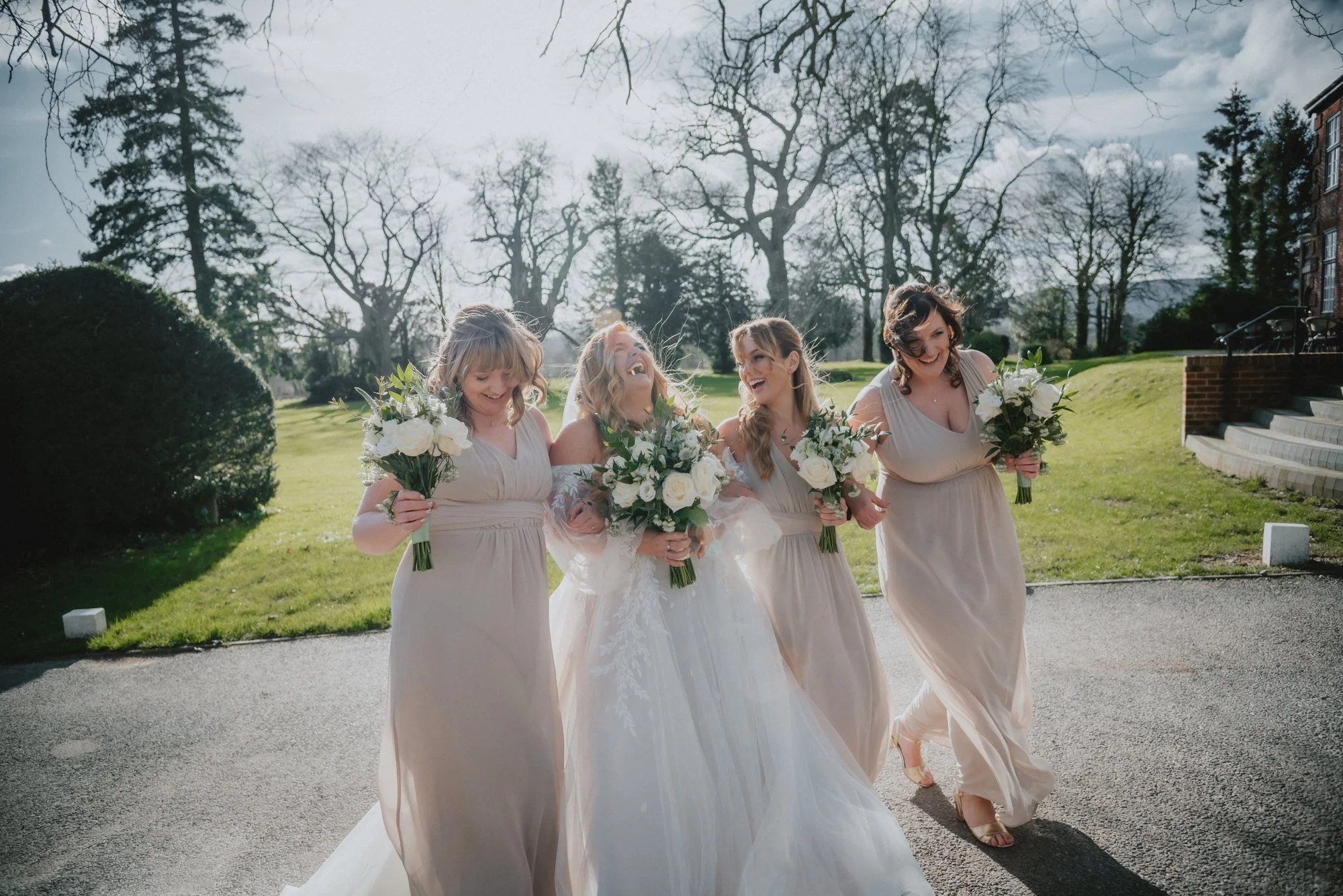 Bridal party of five women in beige dresses walking outdoors on a sunny day, holding bouquets of white roses and greenery.