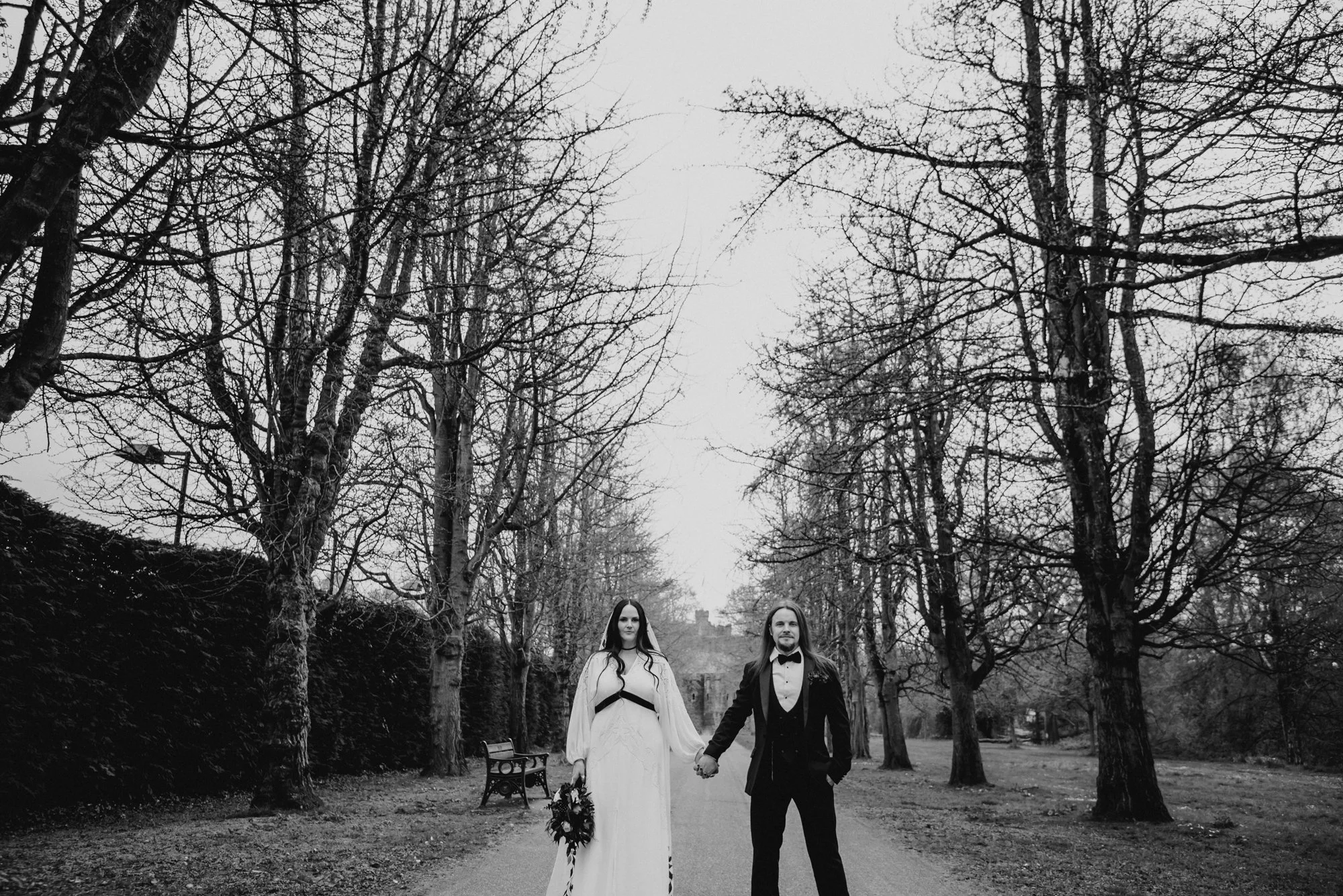 A black and white photo of a couple in wedding attire holding hands in a park with bare trees, a bench, and a path.