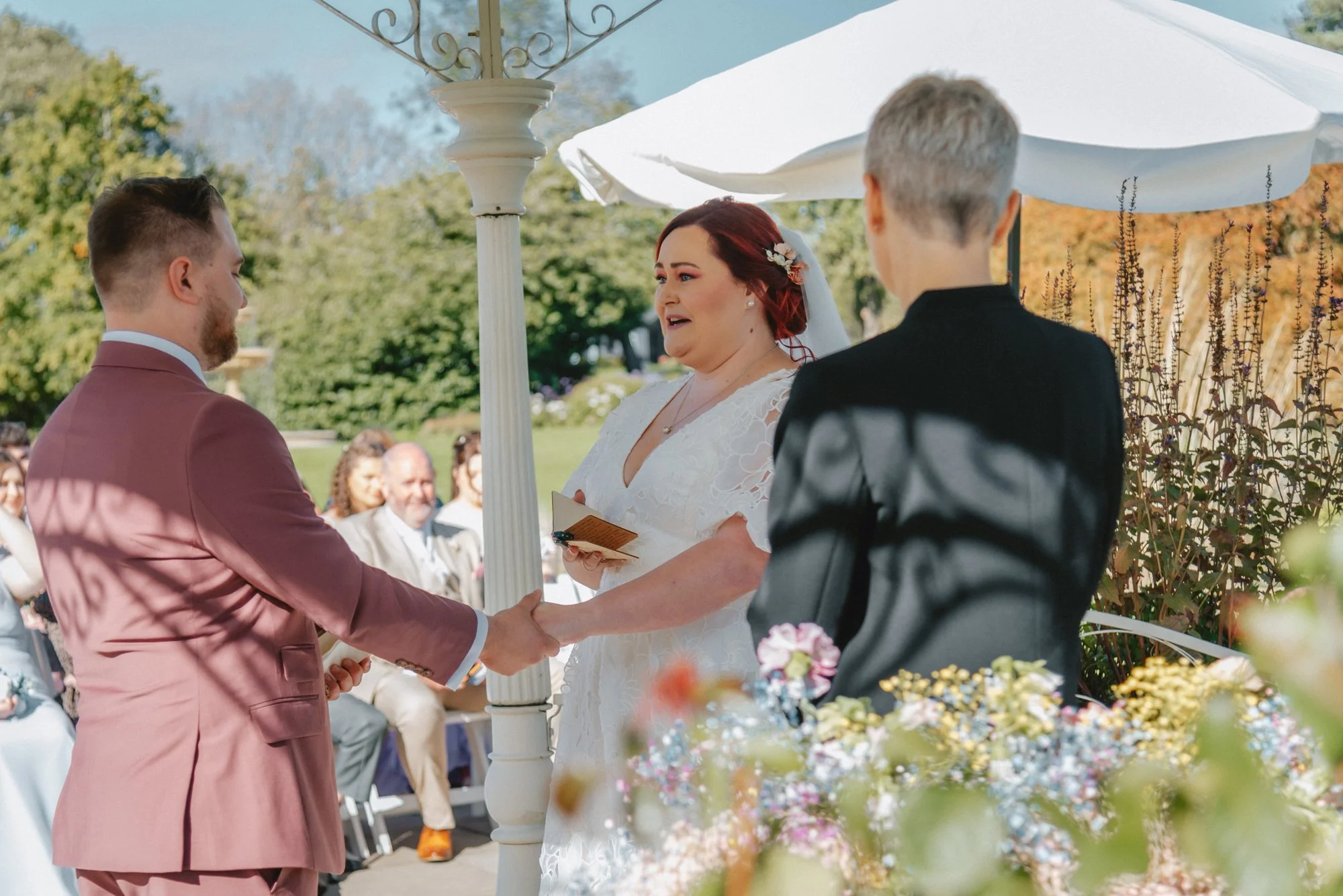 A wedding ceremony with a bride, groom, and officiant outdoors under a gazebo, surrounded by guests, with trees and a blue sky in the background.