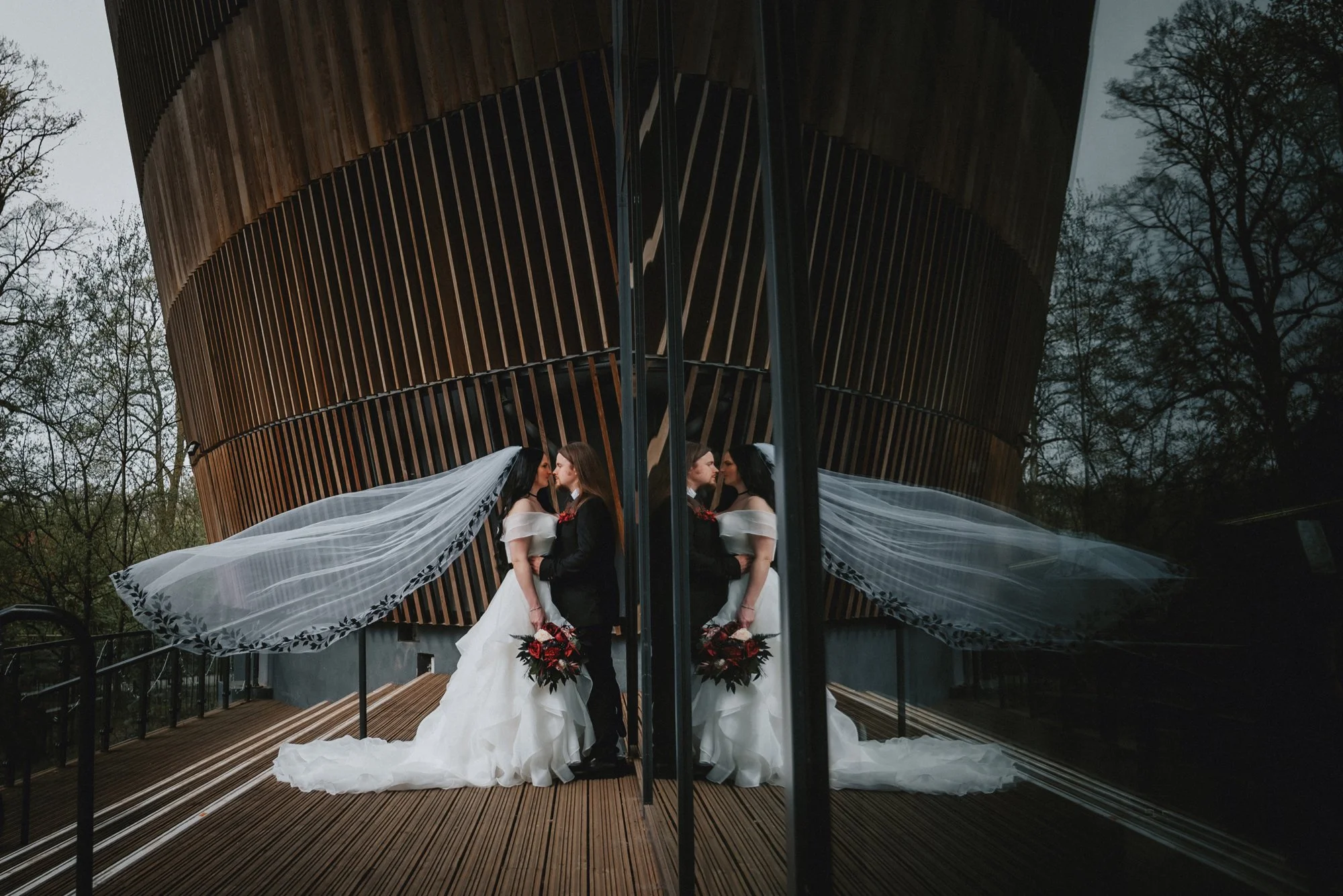 A bride and groom dressed in wedding attire standing close together in front of a reflective glass wall, which creates a mirrored image of them, with a modern wooden building and trees in the background.