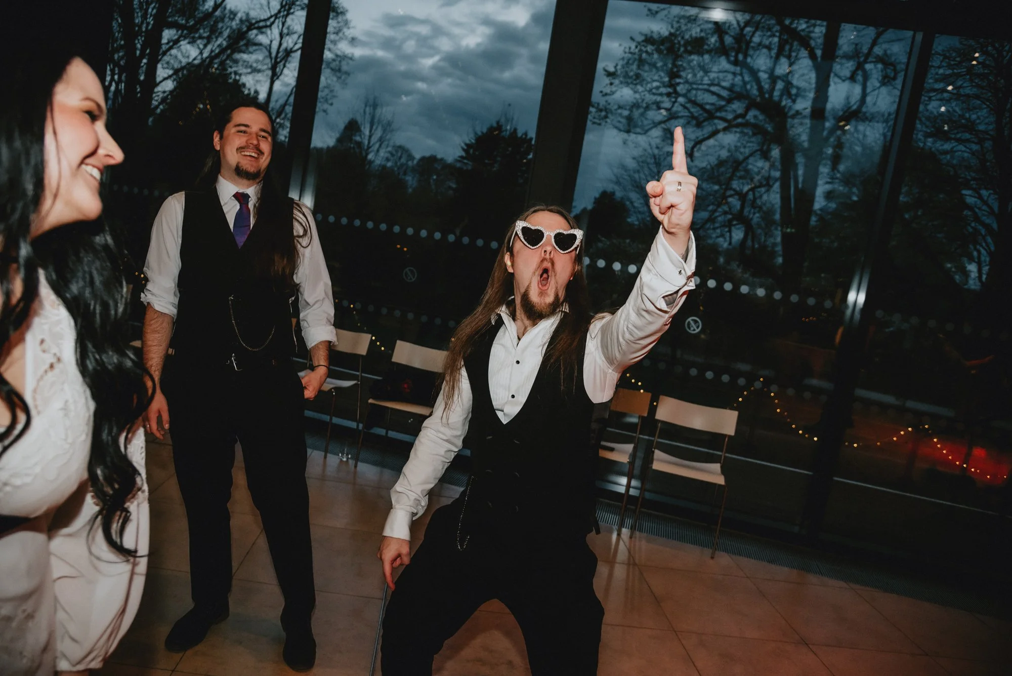 Group of people dancing and having fun at an indoor event during the evening. One man with long hair, sunglasses, a white shirt, and a black vest is striking a pose with his arm raised, while others are smiling and enjoying the moment.