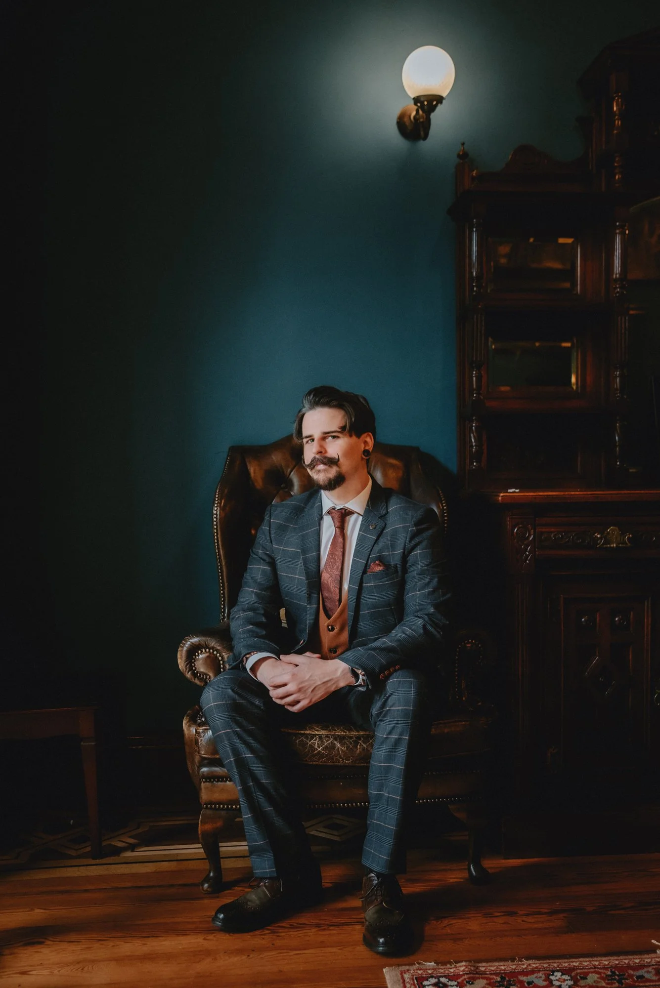 A man in a plaid suit sitting in a vintage leather armchair in a dimly lit room, with dark green walls and wooden furniture.