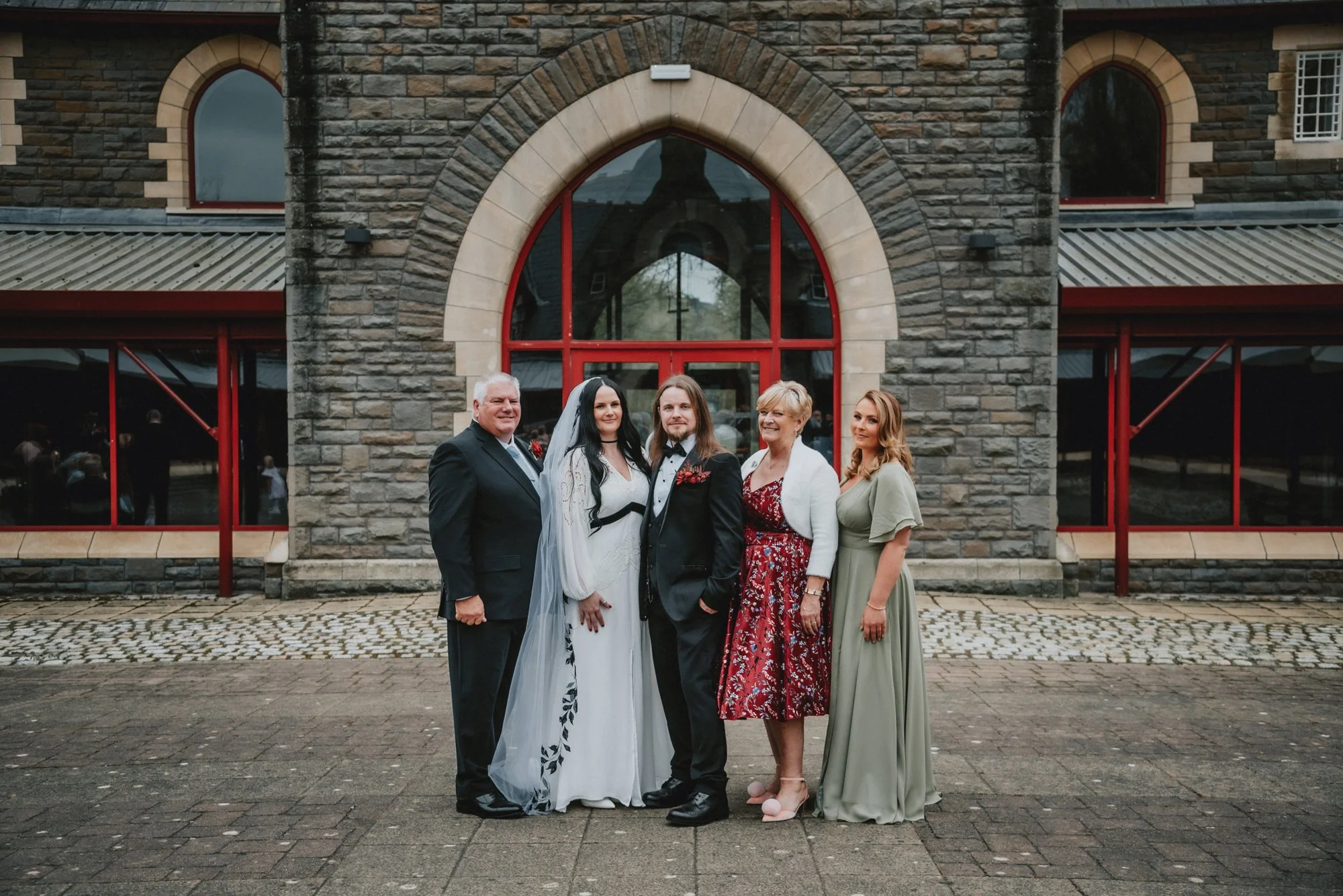 Group of five people standing in front of a stone building with large arched window, dressed in formal and semi-formal attire, during daytime.