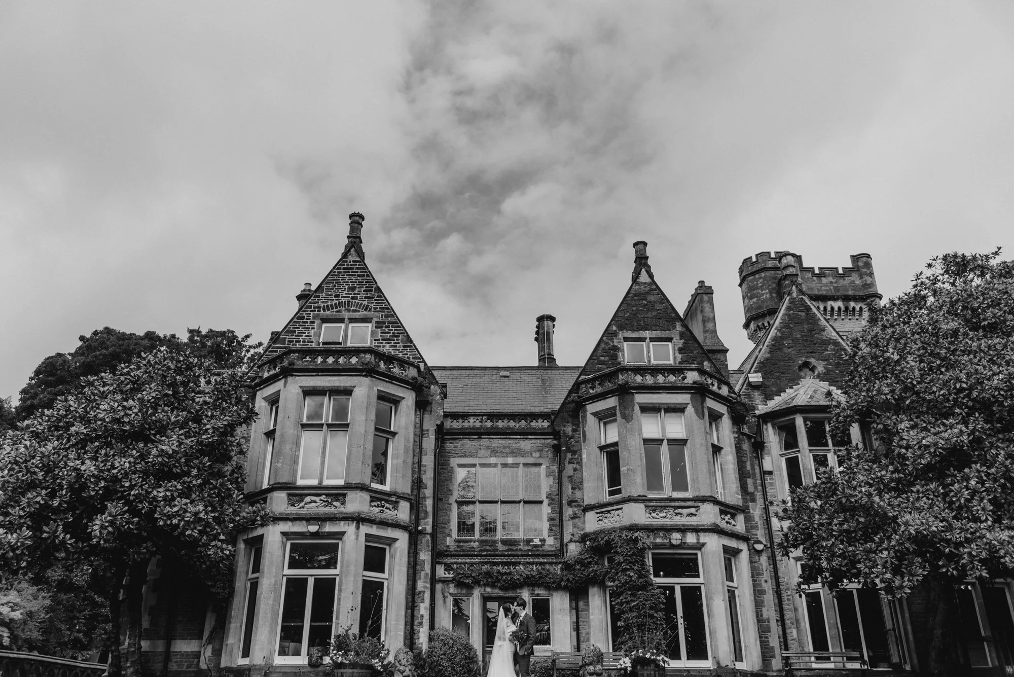 Black and white photograph of a large, historic castle-like building with turrets and bay windows, surrounded by trees. In front of the building, a couple is standing and kissing, with the bride wearing a wedding dress and veil, and the groom in a su