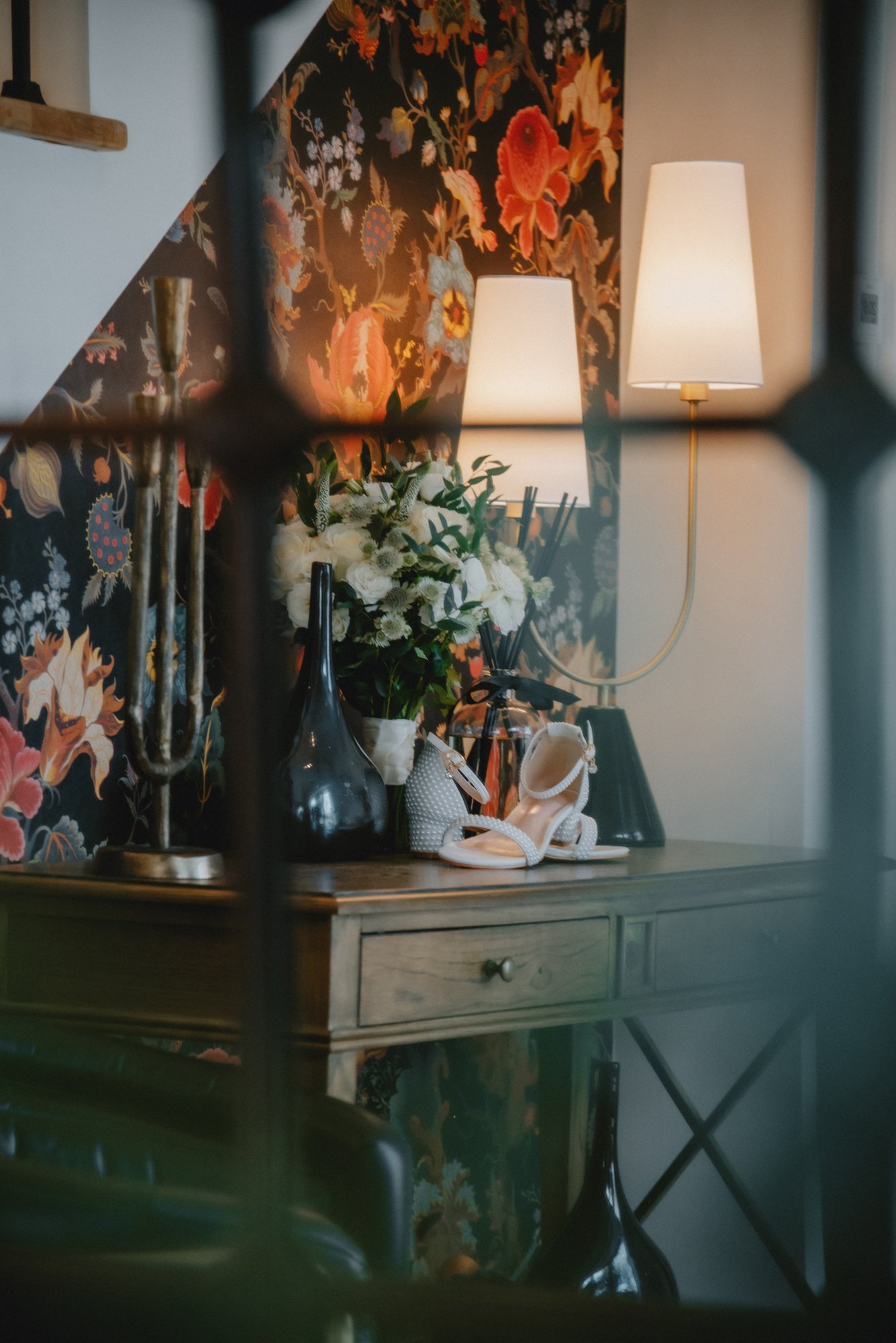 An elegant wooden side table decorated with a black vase, white flowers, jewelry, a pair of beige high-heeled shoes, and two lamps with cream-colored shades. The background features floral wallpaper with large orange and pink flowers.