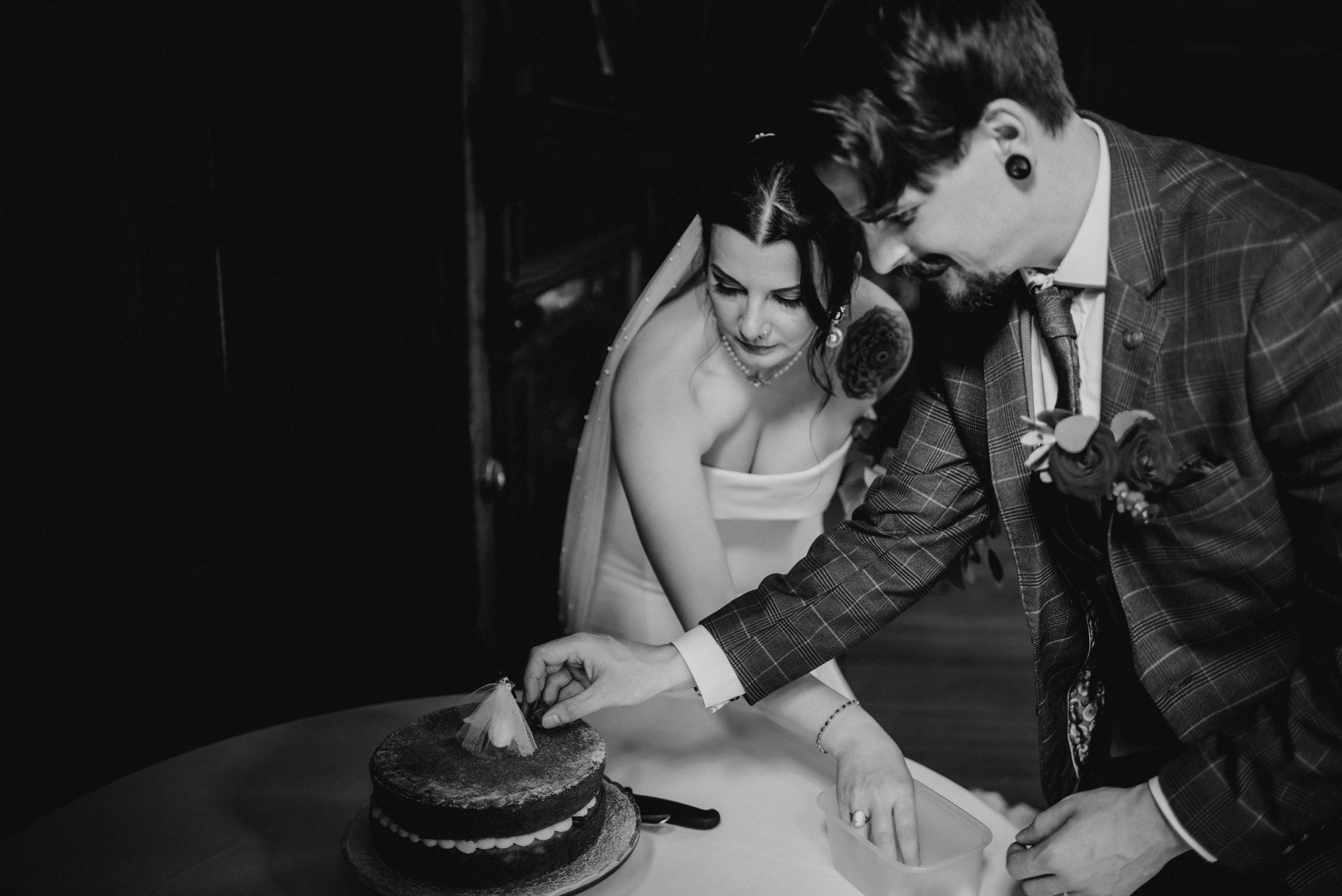 A couple cutting a wedding cake together, with the bride dressed in a strapless gown with tattoos, and the groom in a plaid suit with boutonniere.