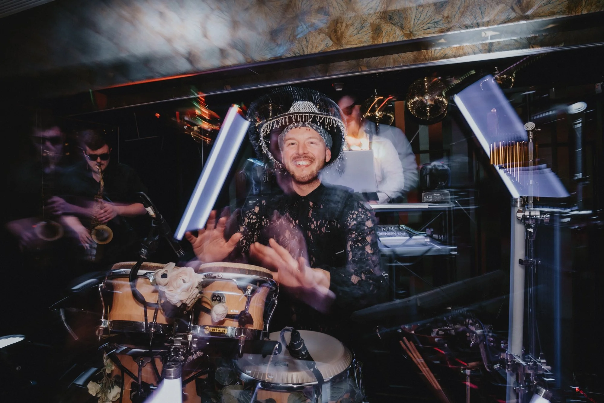 A man with a big smile wearing a decorative hat playing conga drums at a party or celebration, surrounded by musicians and DJ equipment, with disco balls and colorful lights in the background.