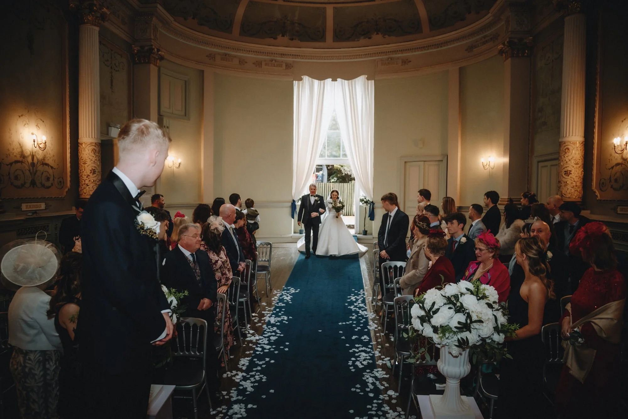 A wedding ceremony in an elegant, historic hall with high ceilings and ornate decorations. The bride and groom walk down the aisle, while guests seated on both sides watch. The aisle is decorated with white flower petals, and there are large floral a