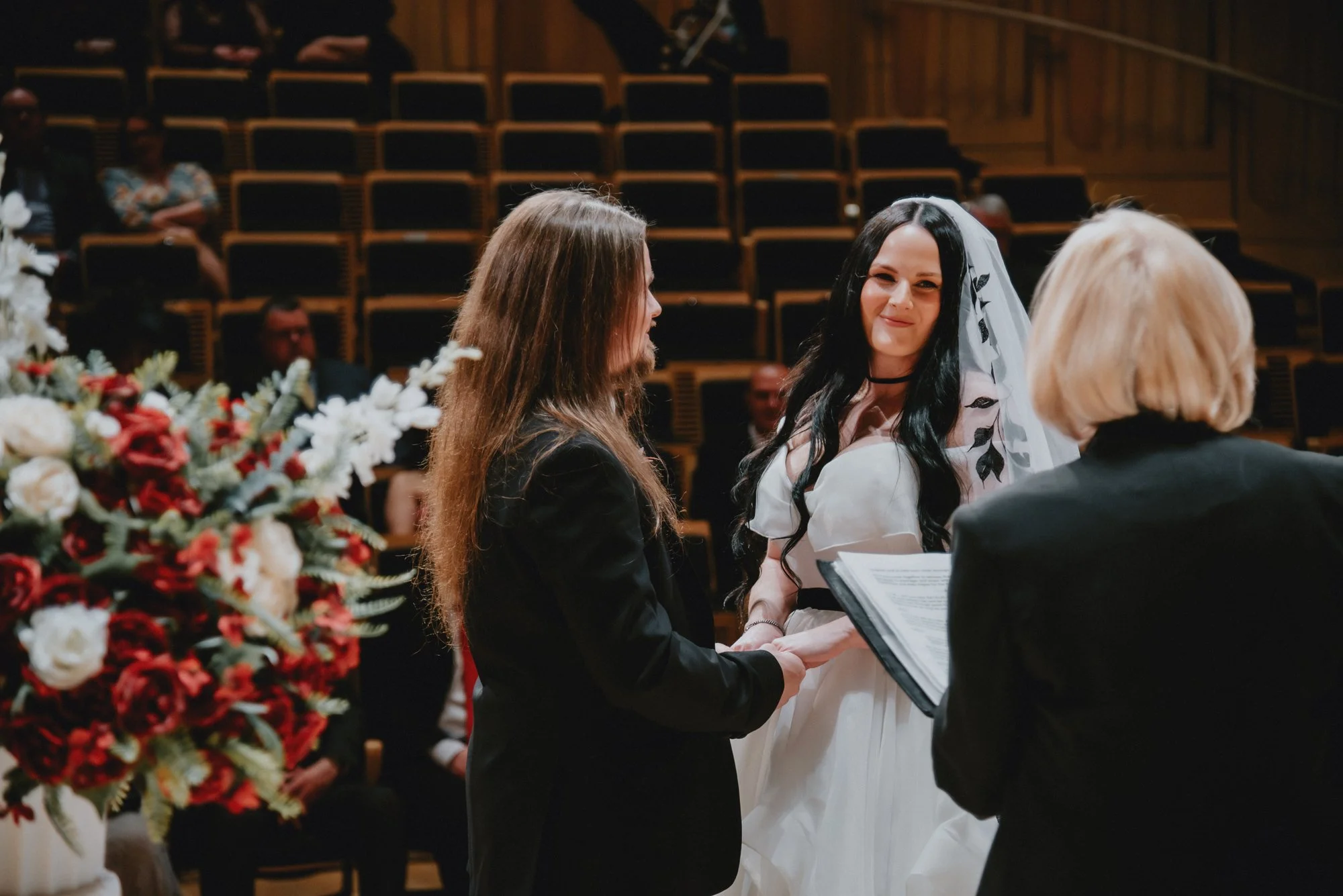 A wedding ceremony with a bride and bride in a formal indoor setting. The bride has long dark hair, a white dress, and a veil, while the other person is dressed in a black suit. An officiant is present, holding a book, and there are guests seated in 