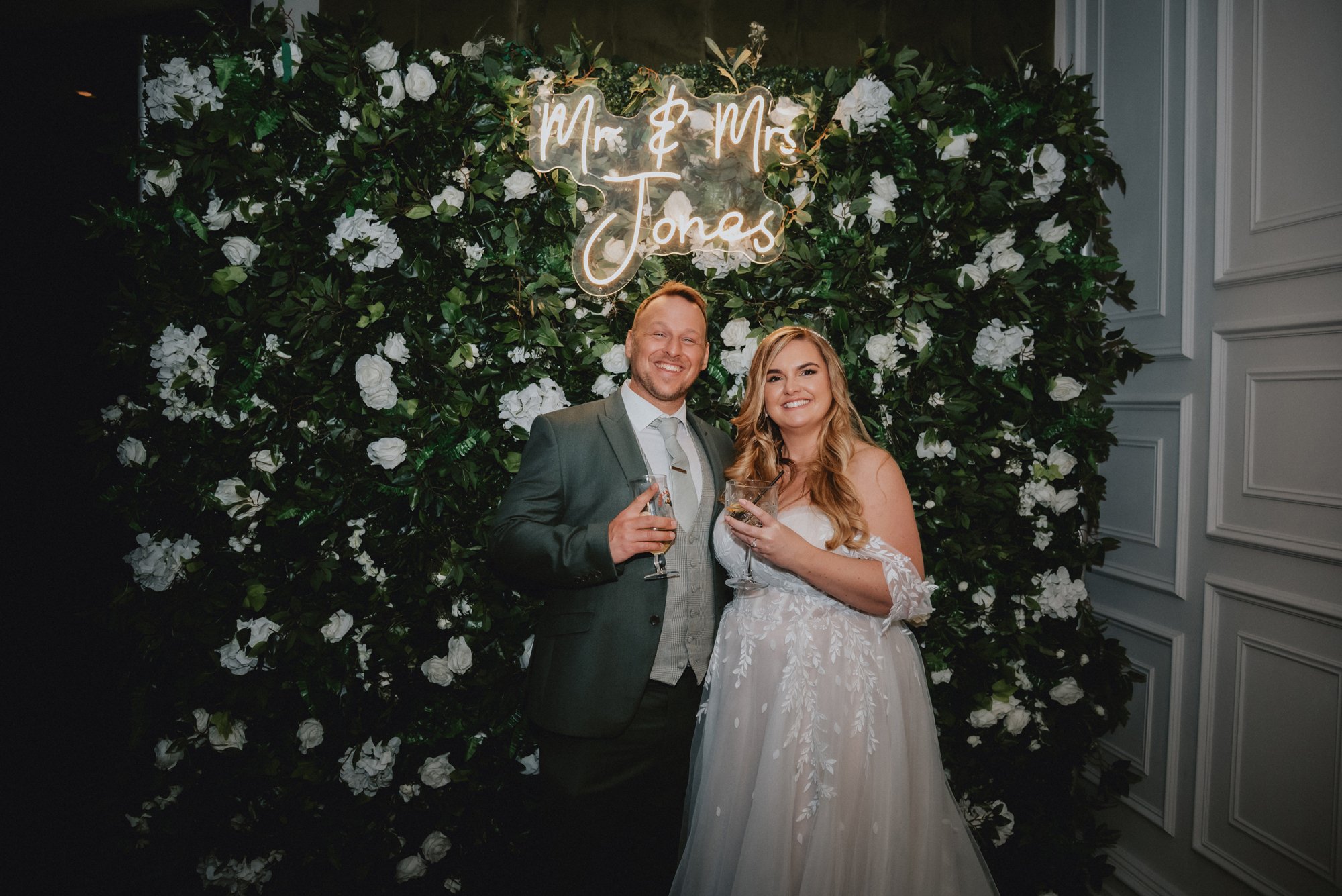 A newlywed couple at their wedding reception, standing in front of a large green floral backdrop adorned with white flowers and a neon sign reading 'Mr & Mrs Jonas'. The groom is wearing a gray suit with a white shirt and a gray tie, holding a drink,
