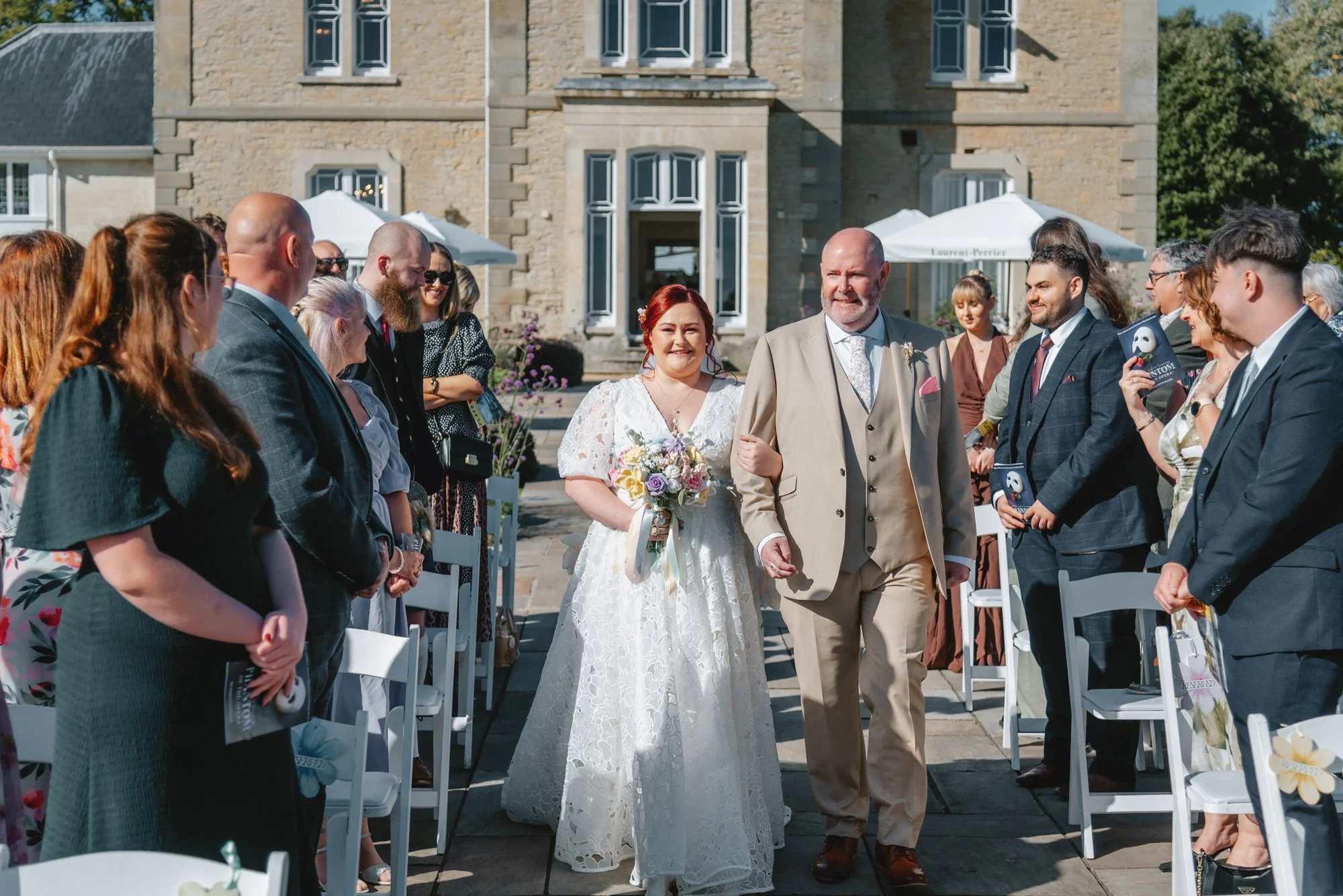 Bride walking arm-in-arm with her father during a wedding ceremony outside a historic building, surrounded by seated and standing guests.