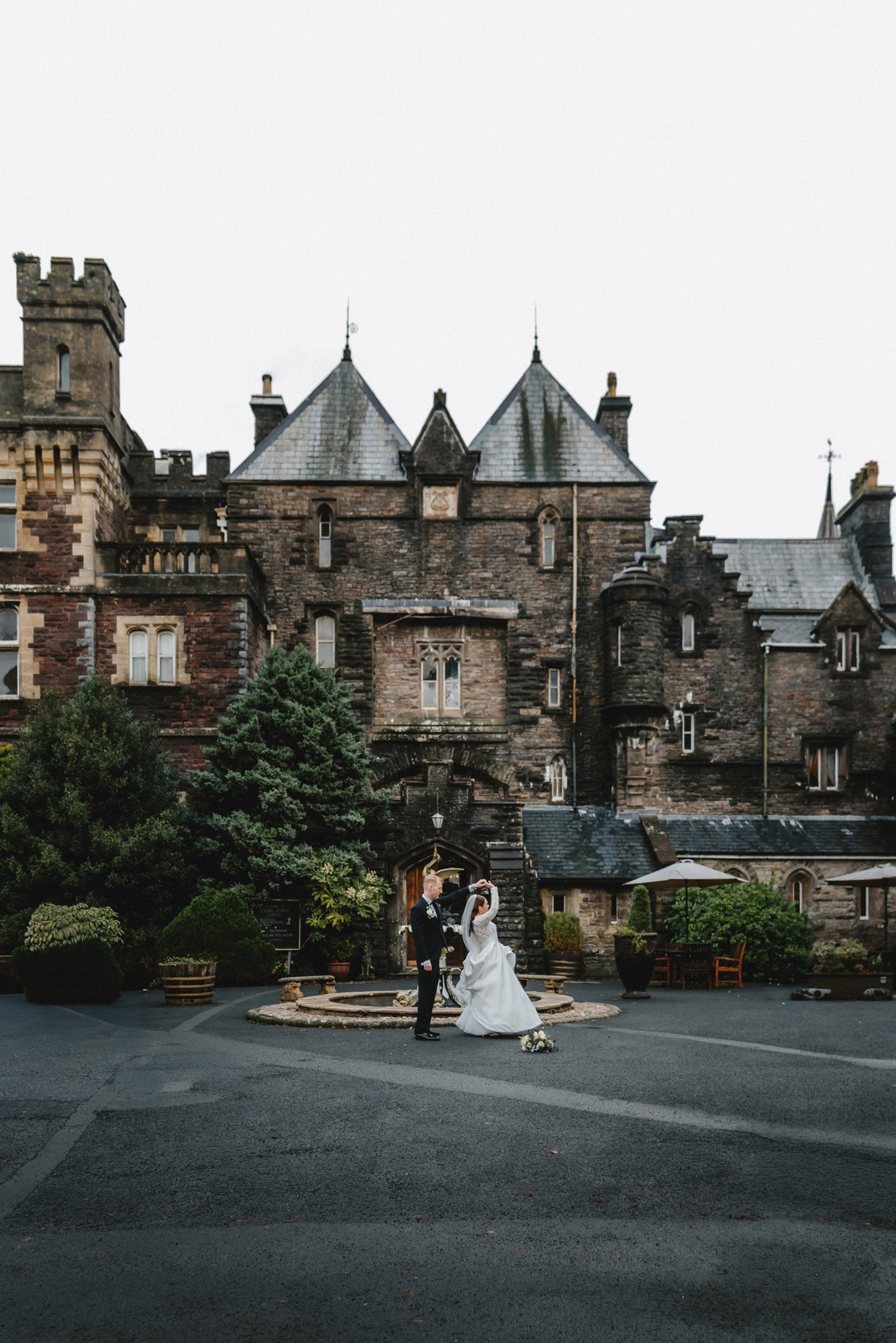A bride and groom dancing in front of a historic stone castle on their wedding day, with the bride in a white gown and the groom in a black suit, surrounded by trees and outdoor furniture.