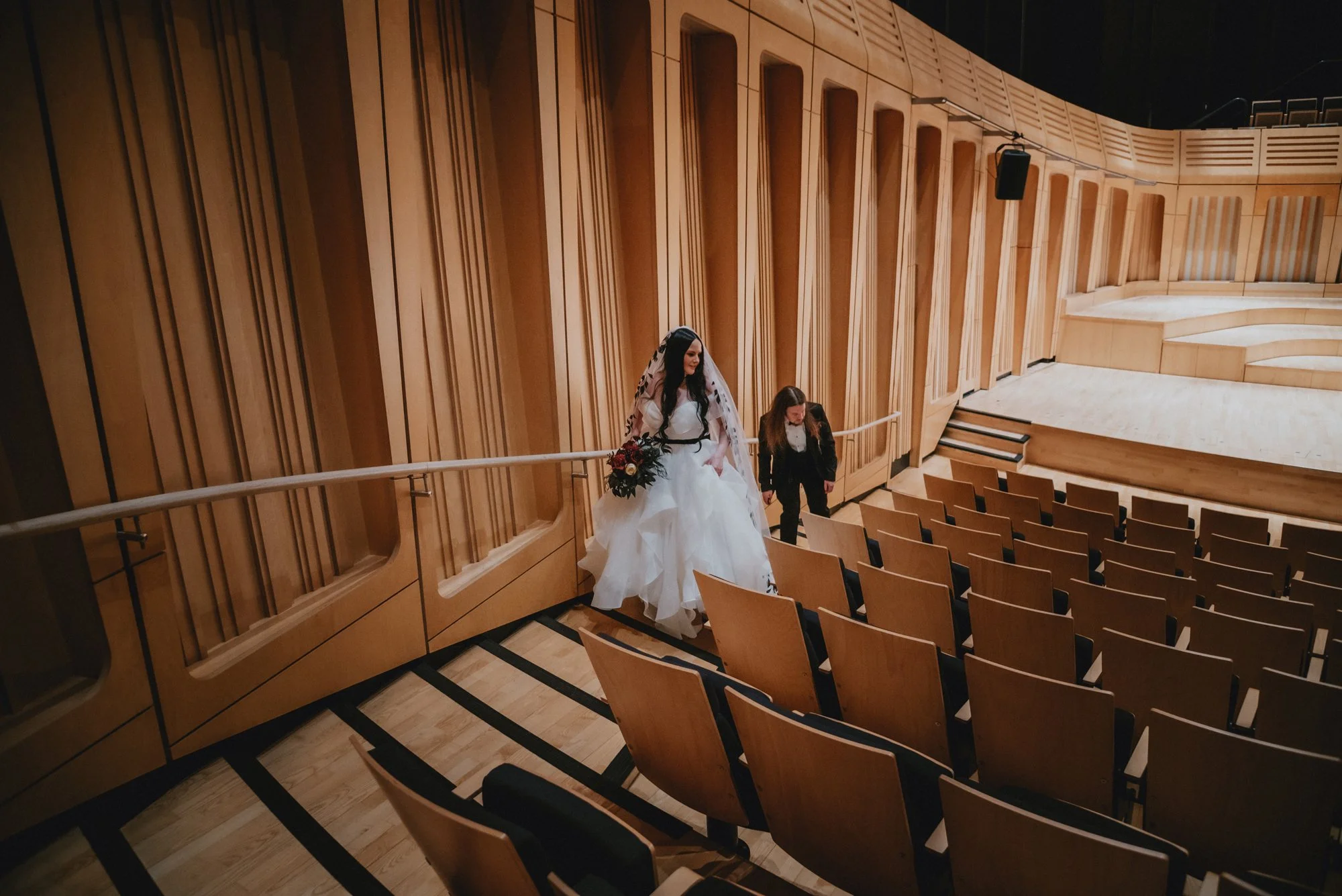 A bride in a white wedding gown and veil, holding a bouquet, walking down the aisle of an auditorium with wooden walls and empty seats, accompanied by a woman in black attire, possibly a wedding planner or assistant.