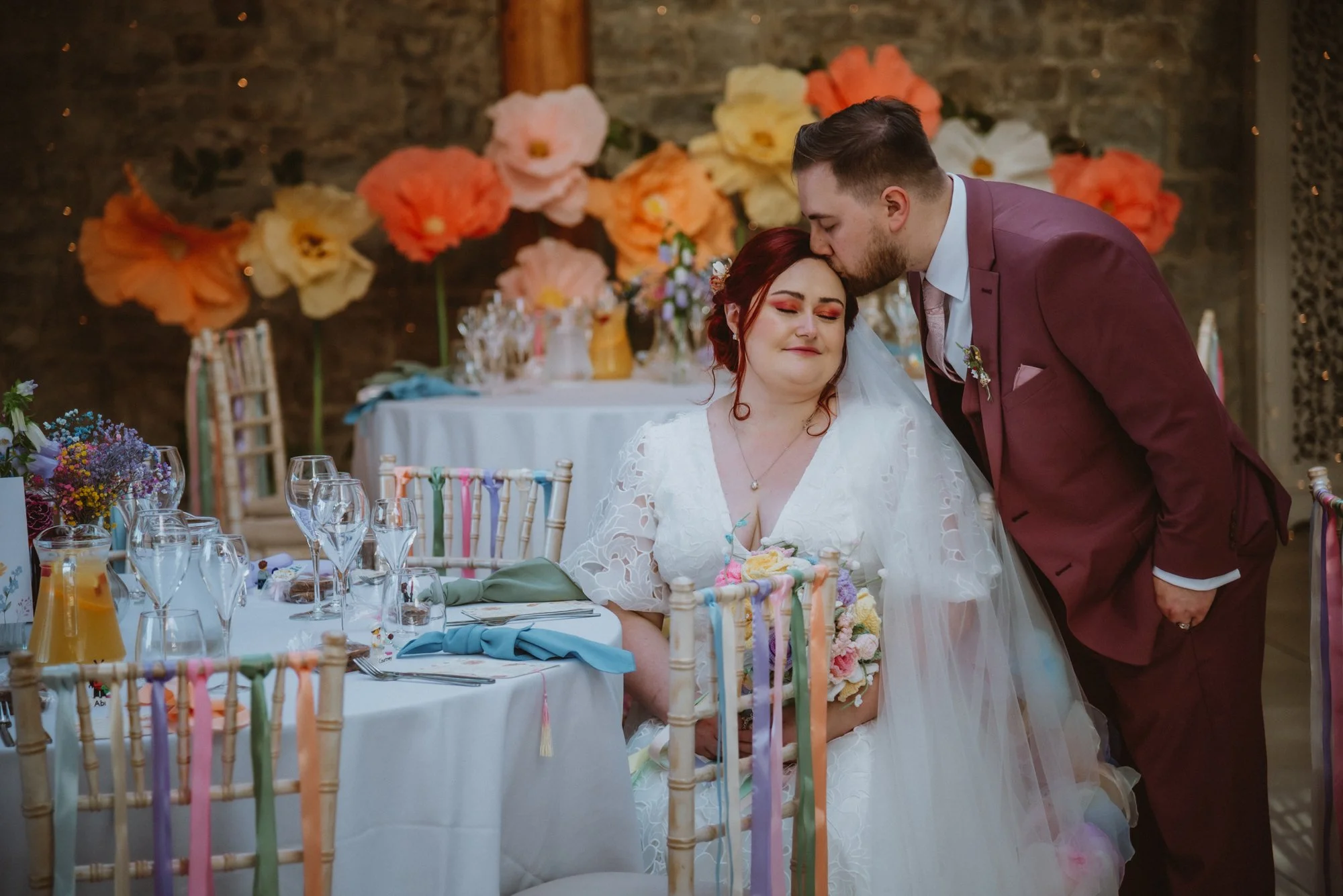 A bride with red hair and a white dress sitting at a decorated wedding table with colorful ribbons, holding a bouquet, and a groom in a burgundy suit kissing her forehead at a wedding reception with large paper flowers on the wall in the background.