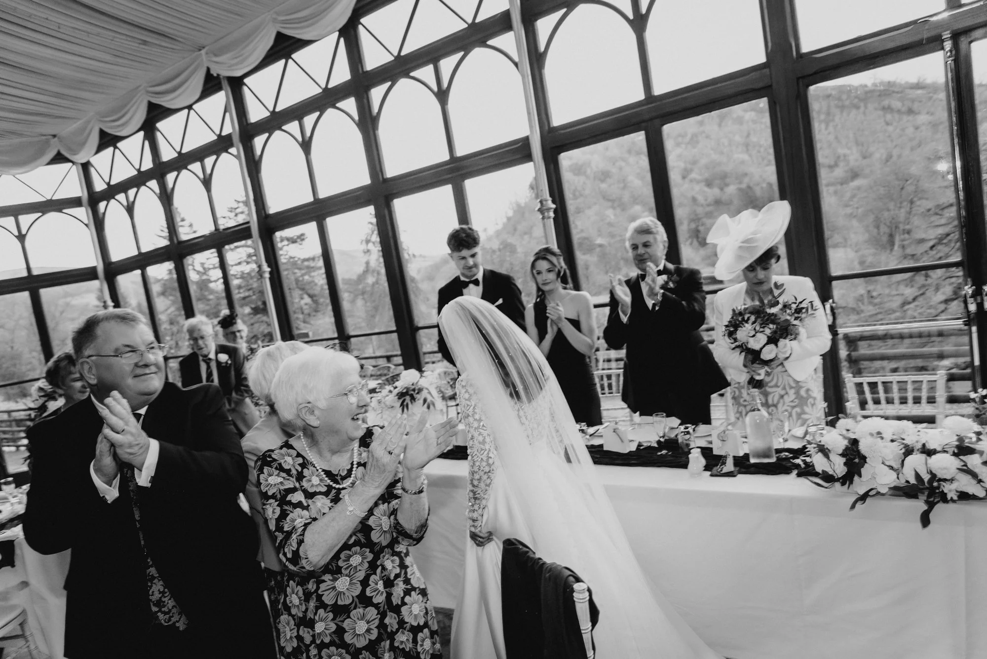 A bride with a veil and white gown leans forward to greet an older woman, surrounded by clapping guests in a glass-enclosed wedding venue