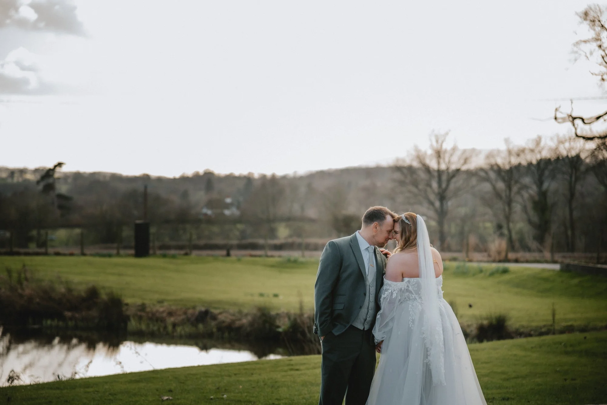 A bride and groom sharing a tender moment with foreheads touching outdoors on a grassy area near a pond, trees, and rolling hills at sunset.