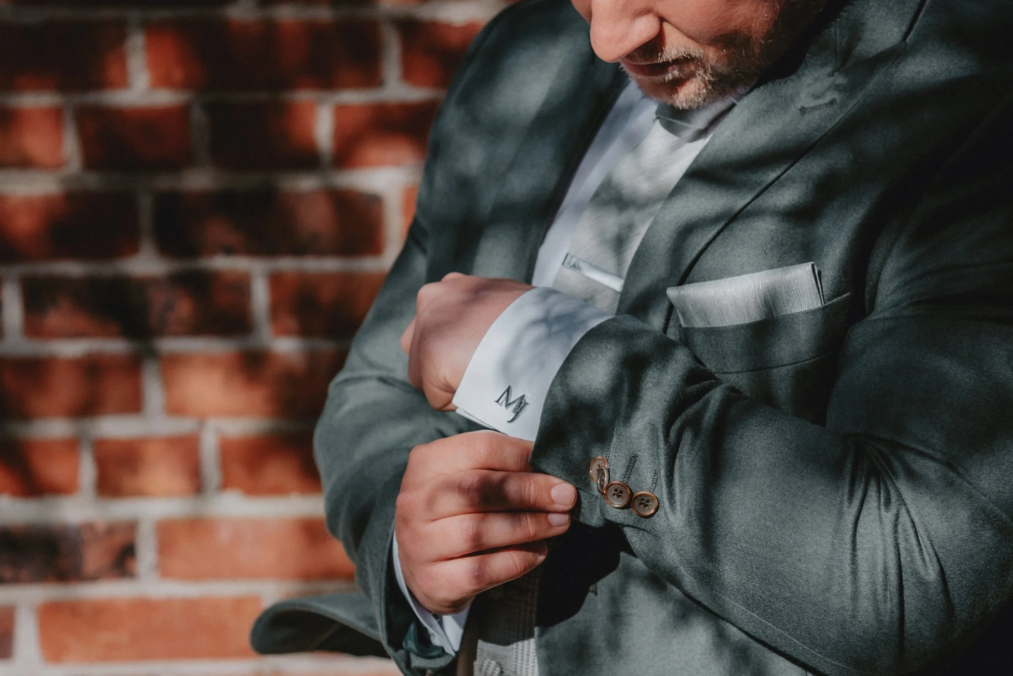 A man wearing a gray suit adjusting his shirt cuff in front of a brick wall.