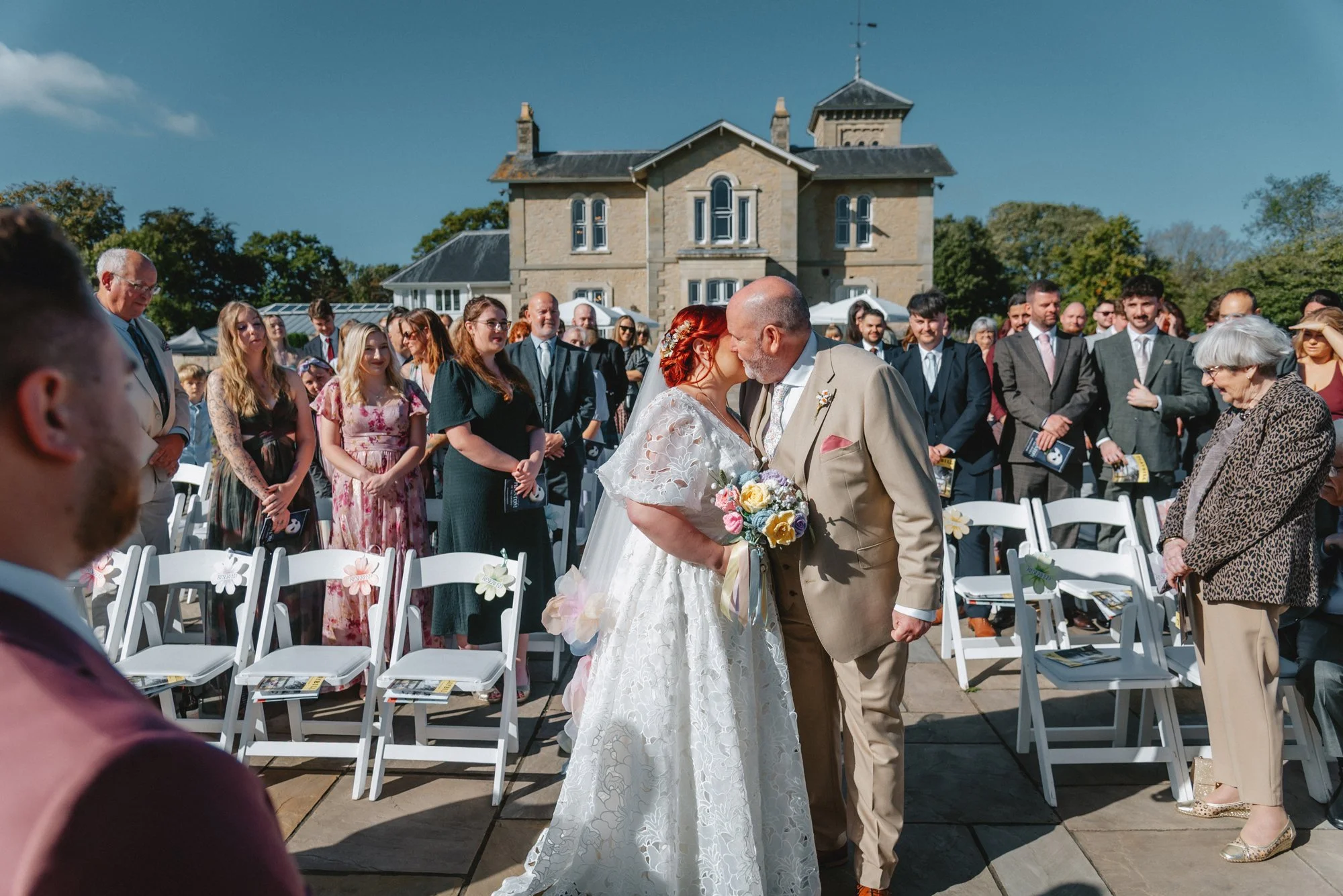 Wedding ceremony outdoors with bride and groom kissing, guests standing and sitting around, large historic house in the background on a sunny day.