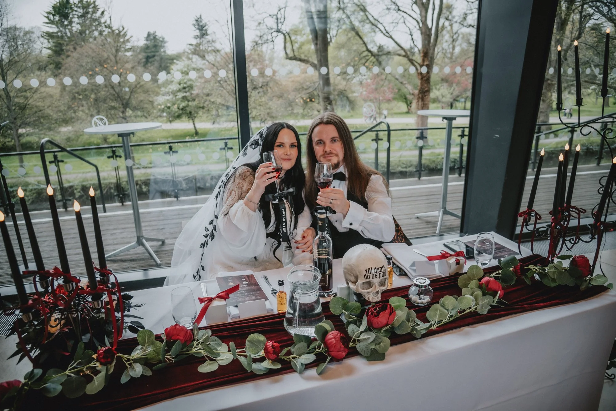 Couple dressed in Halloween costumes sitting at a decorated dinner table, holding glasses of red wine, with a backdrop of trees and greenery outside the window