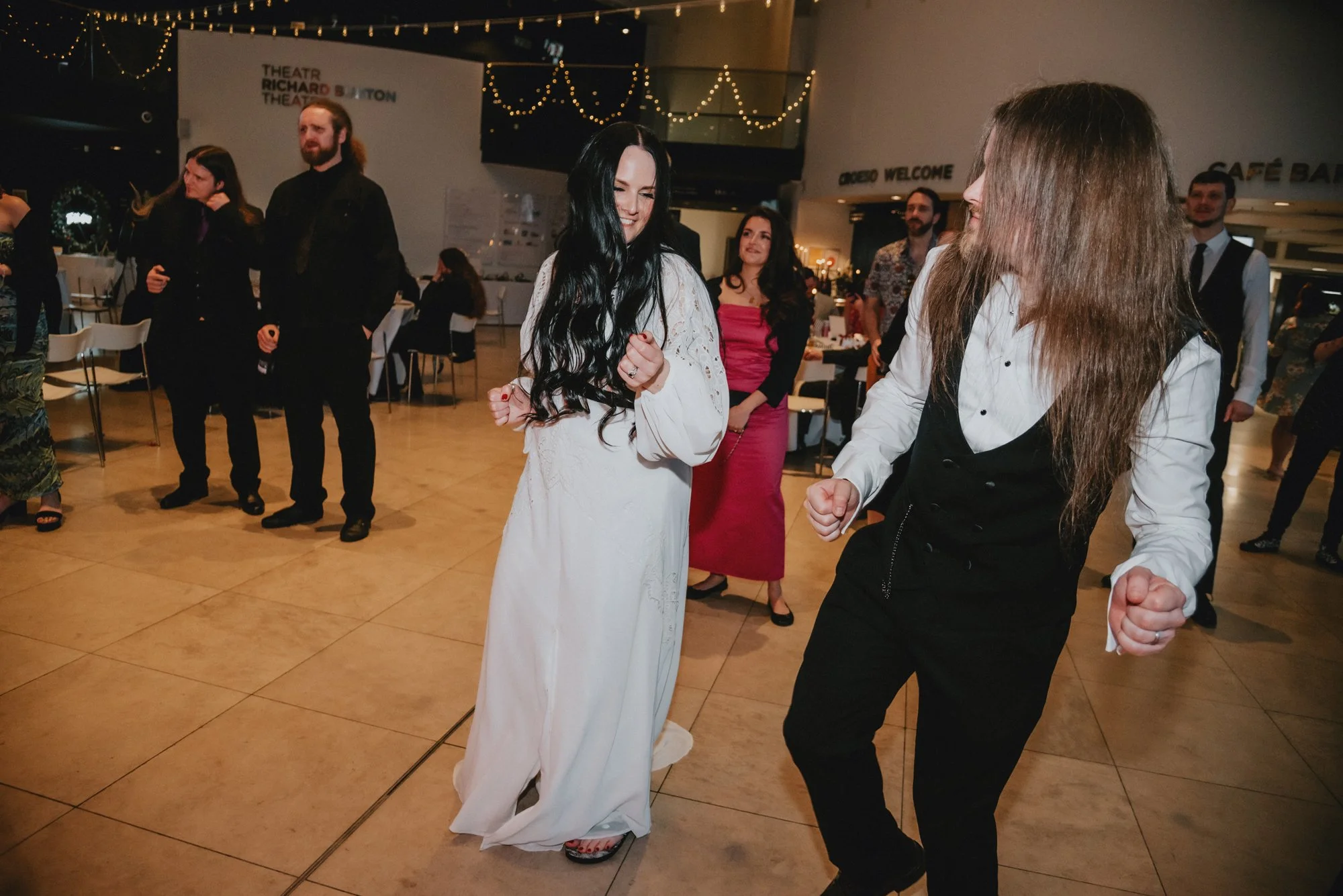 Two women dancing and smiling at a wedding reception with other guests in the background.