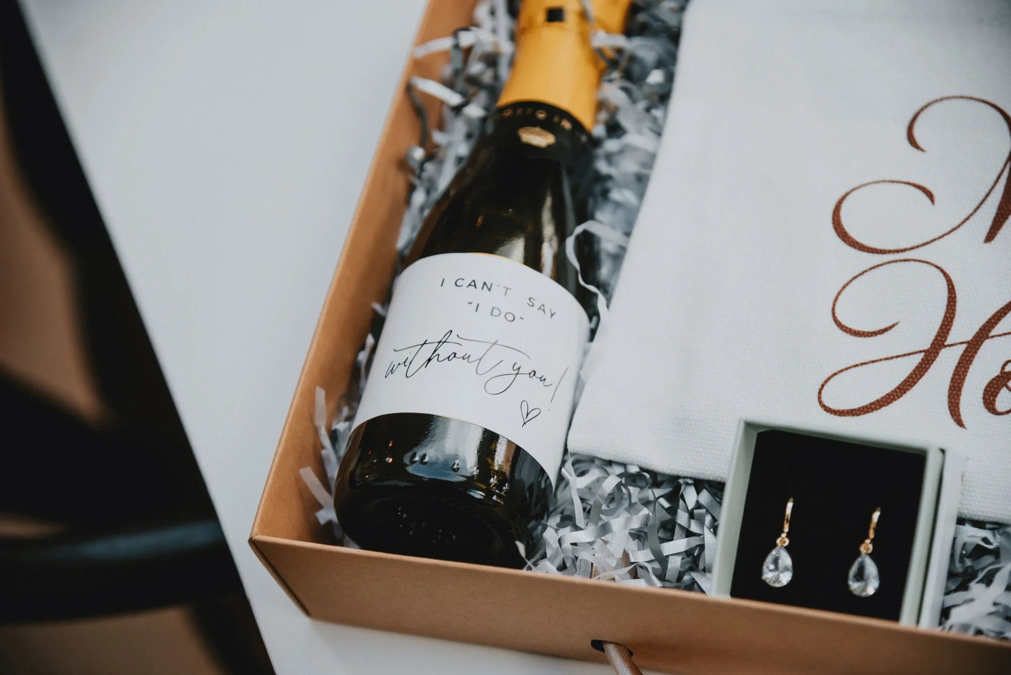A gift box containing a small bottle with a white label that reads 'I can't say "I do" without you!', a pair of gold earrings with teardrop-shaped stones in a small white box, and a white canvas with elegant script lettering.